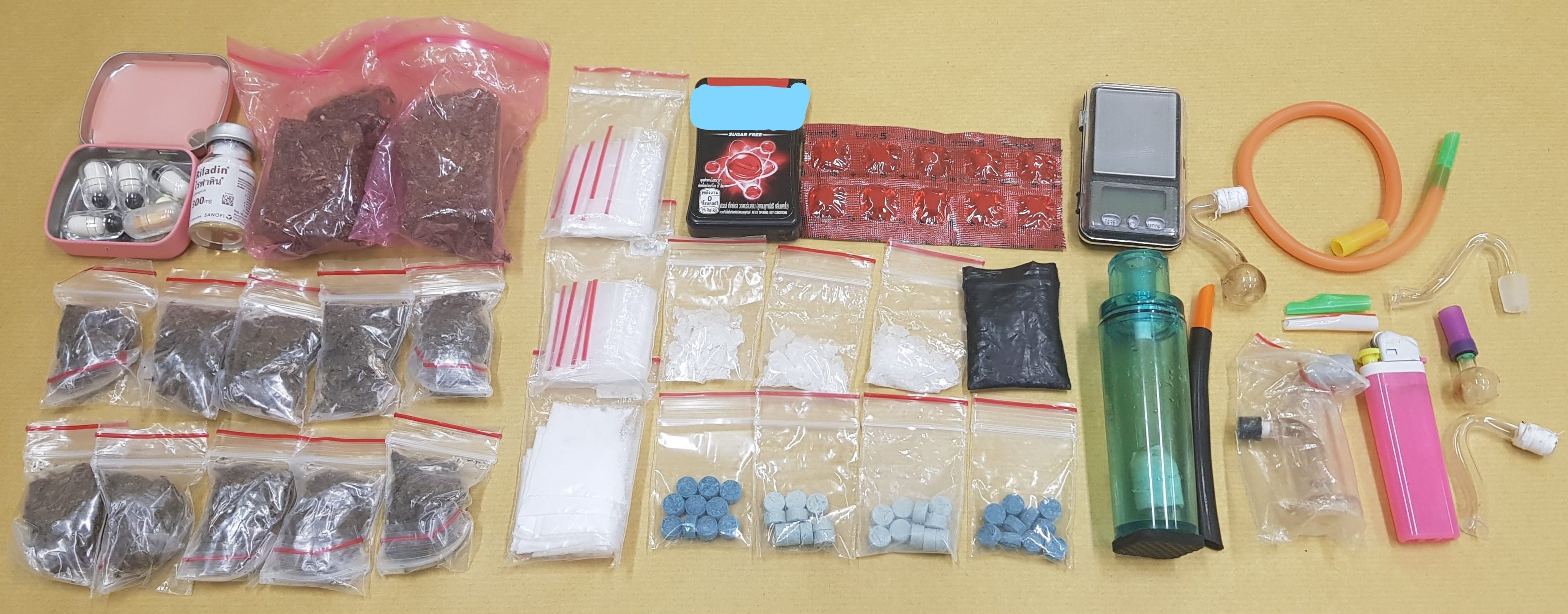 Various bagged substances, pills, scale, pipes, and lighter on a light brown surface.