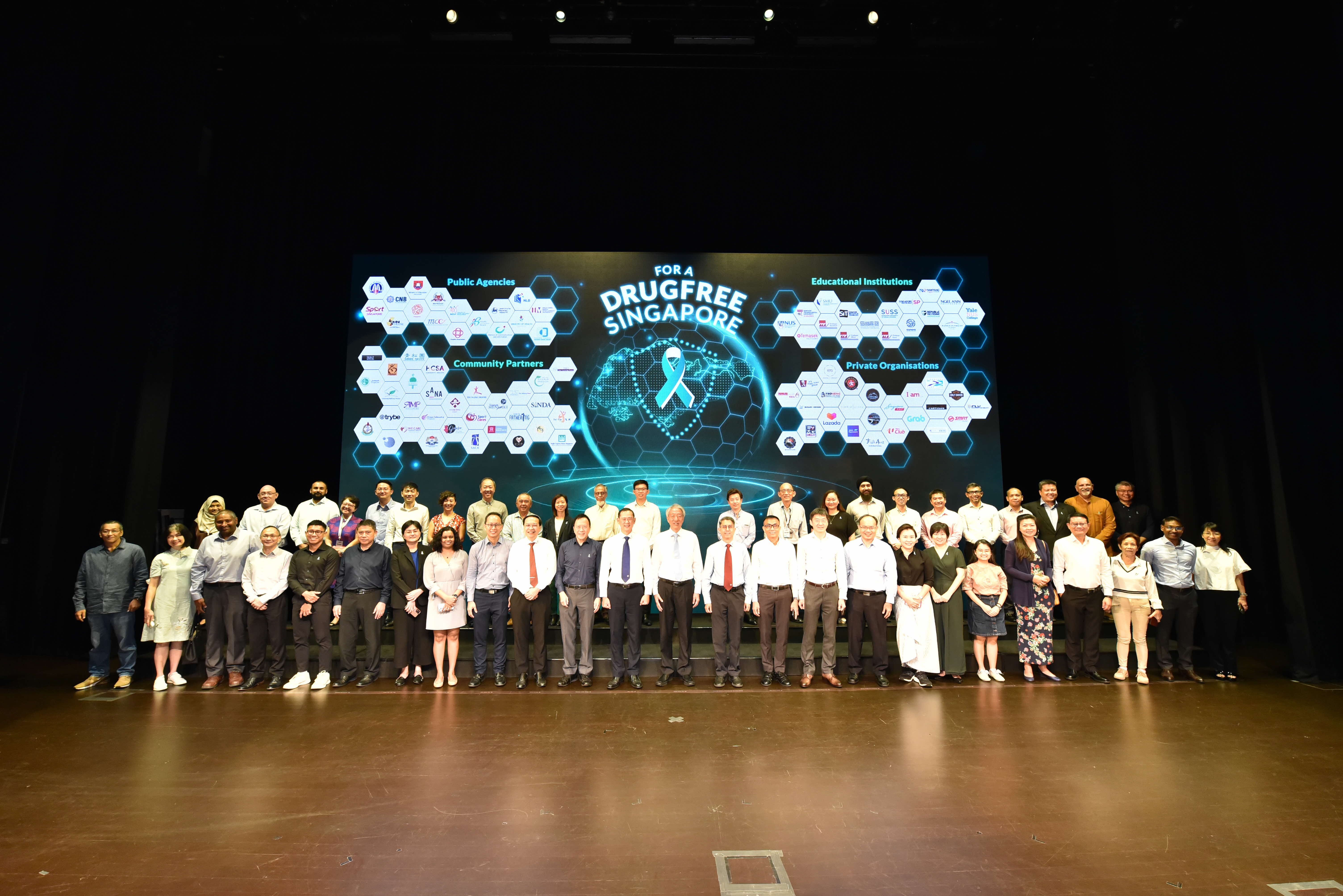 Group of people on stage, with "For A Drugfree Singapore" logo and partner organizations on a large screen behind them.