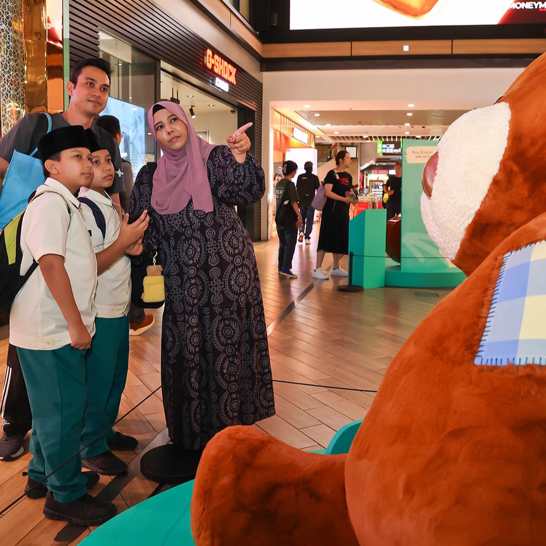 A family with two schoolchildren listens as a woman points toward a large teddy bear display in a shopping mall.