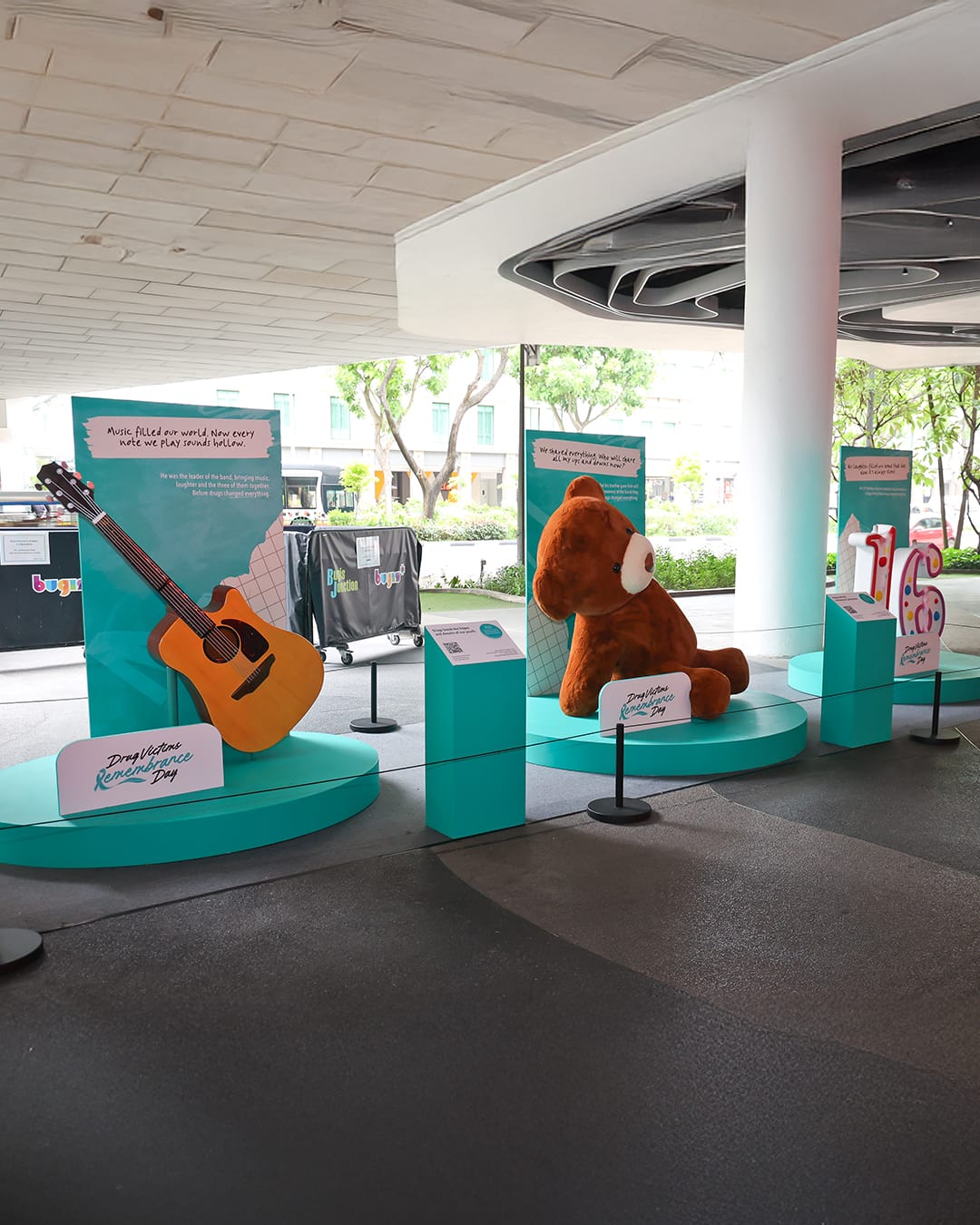 Outdoor exhibit showing a guitar, large teddy bear, and number sculpture for Drug Victims Remembrance Day.