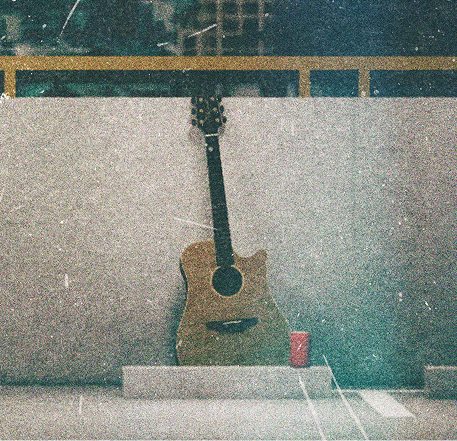 Acoustic guitar leaning on wall with a red cup beside it, in a grainy, vintage-styled photo.