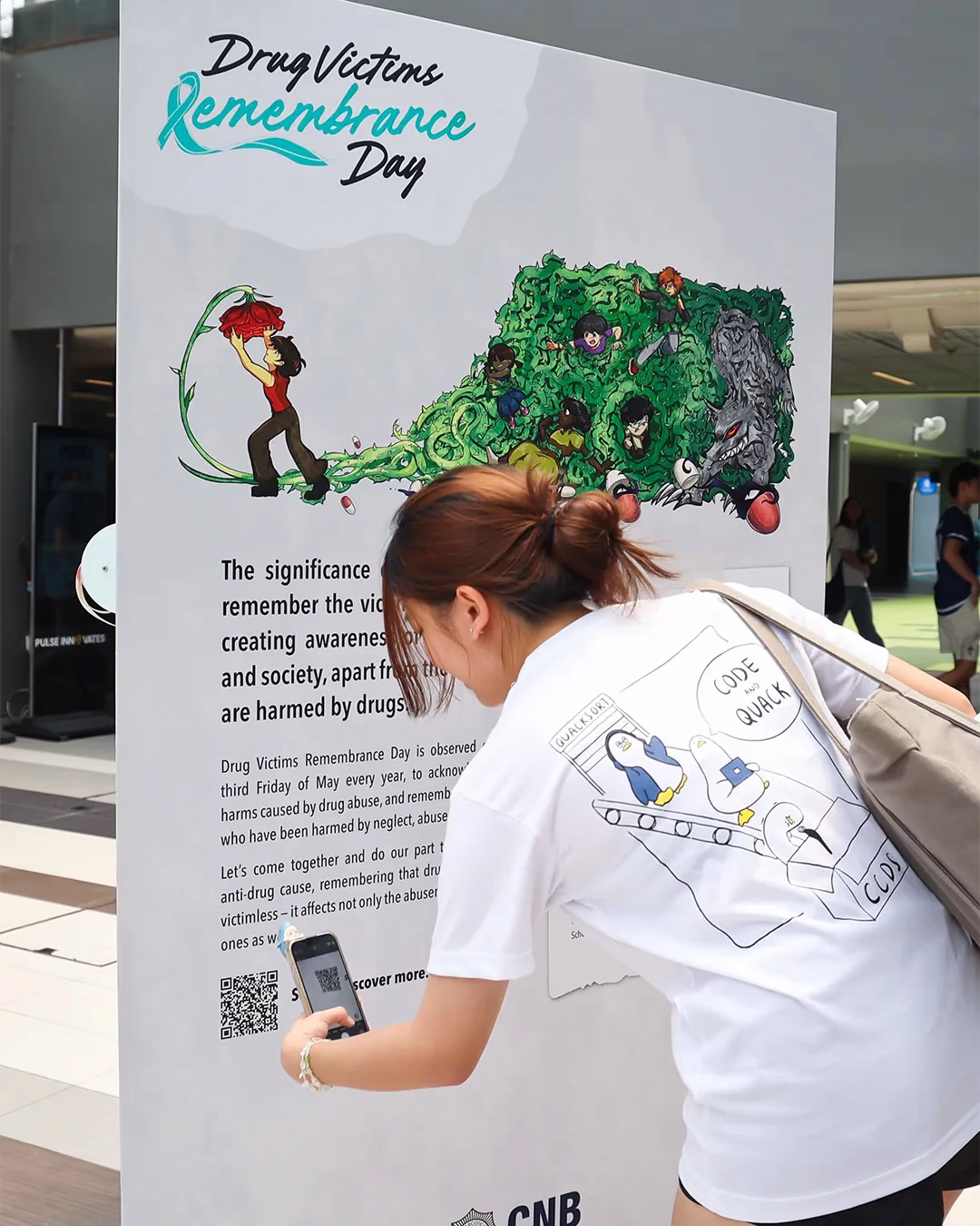 A woman scans a QR code on a Drug Victims Remembrance Day information board with illustrated artwork.