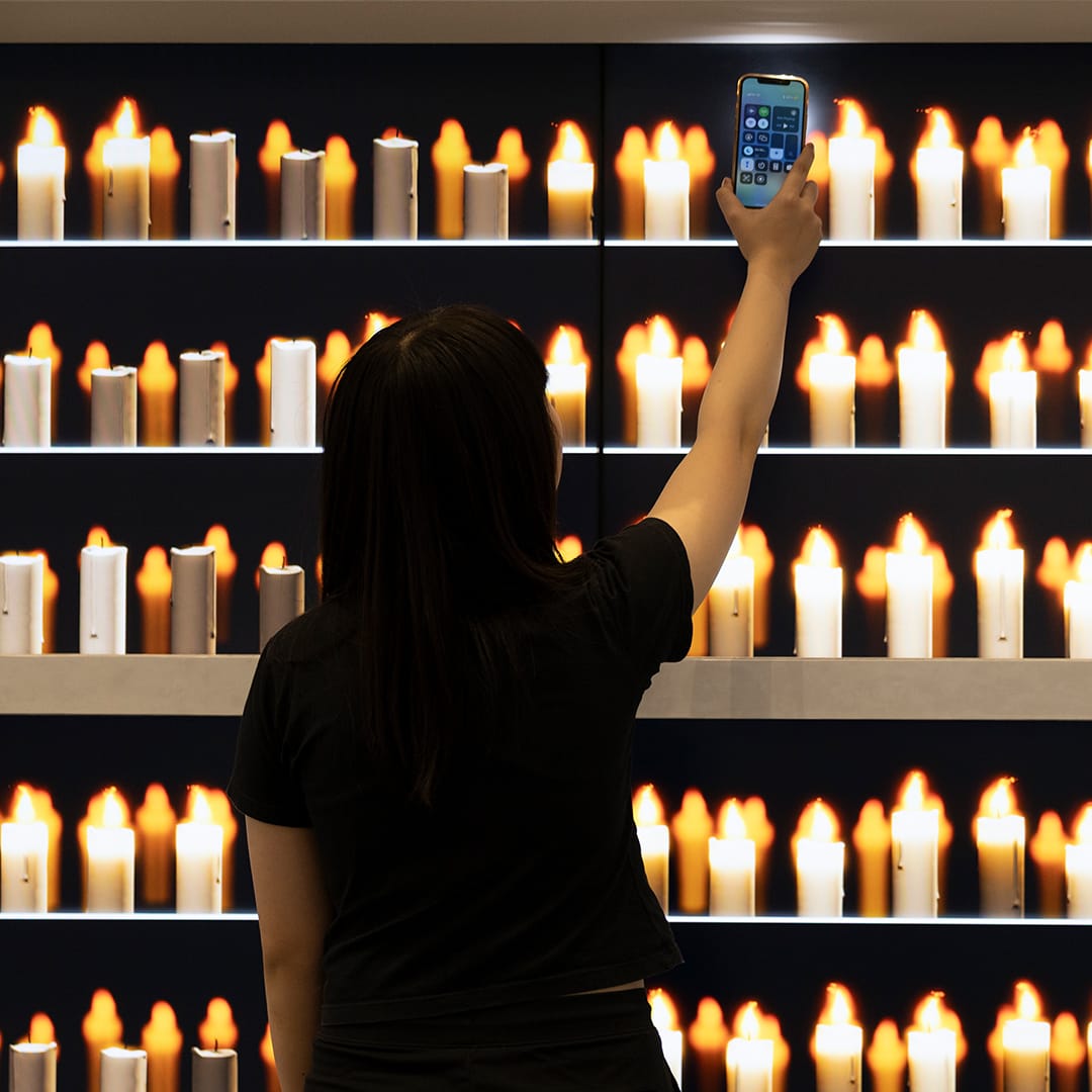 Visitor holding up a phone before a wall of glowing digital candles.