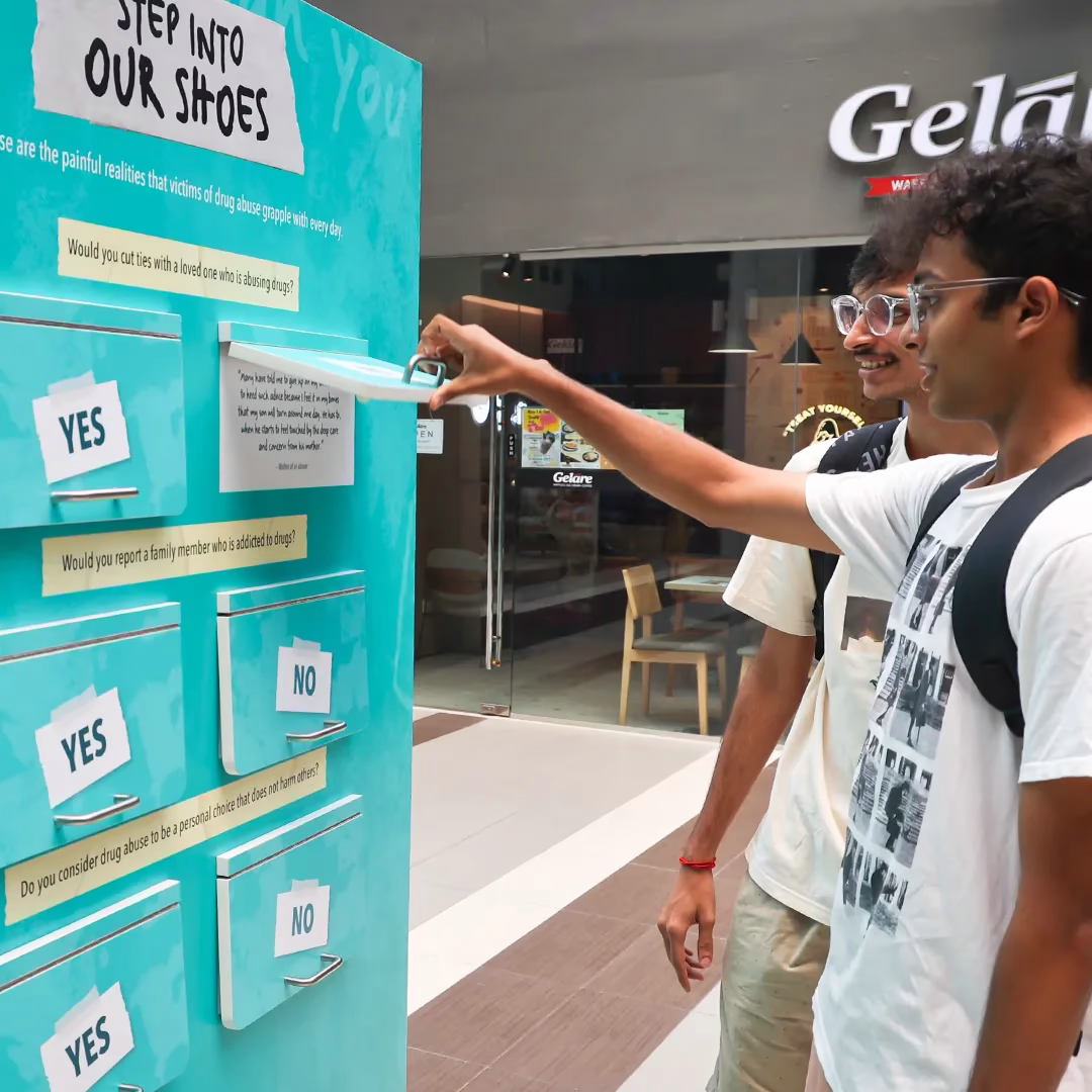 Two young men interact with a “Step Into Our Shoes” display, opening a panel with questions about drug abuse.
