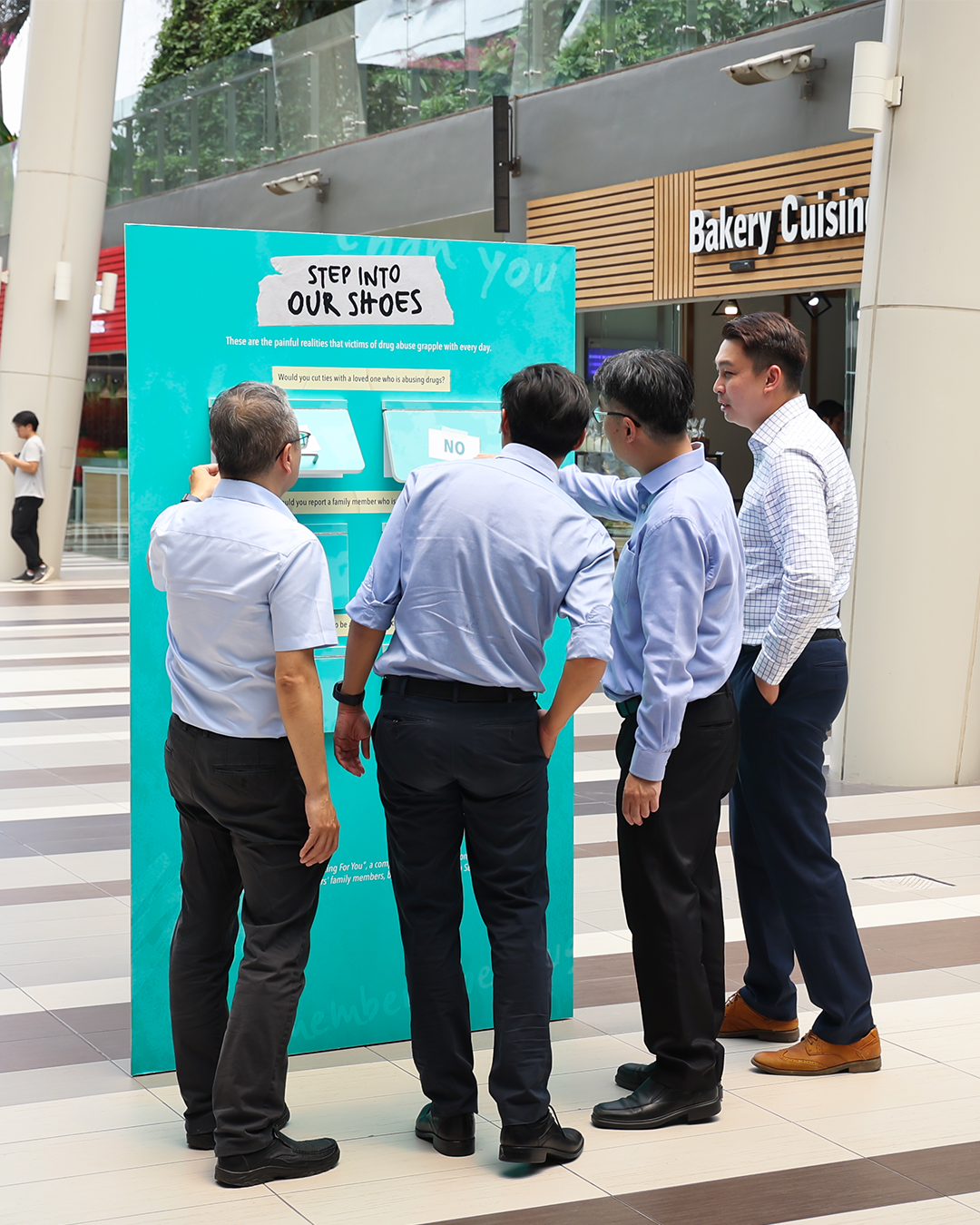 Five men in office attire engage with a “Step Into Our Shoes” display, discussing its questions.