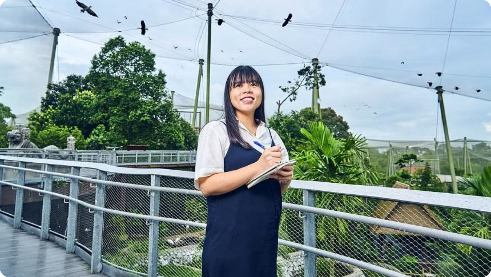A person stands on a mesh-fenced walkway holding a notepad, surrounded by trees and flying birds.