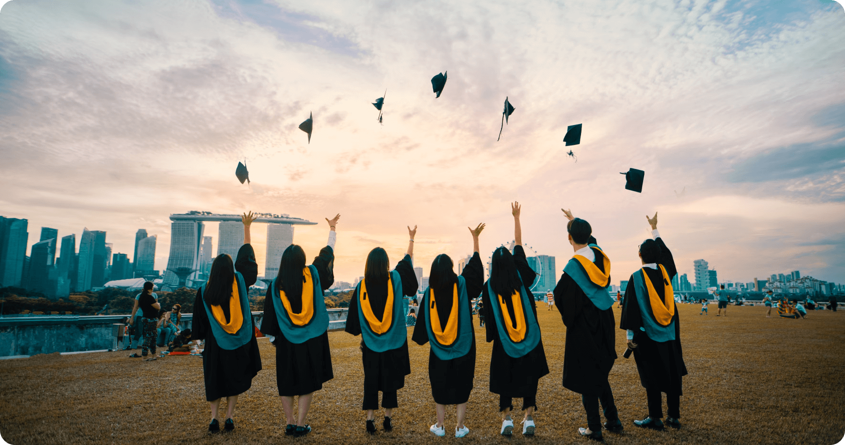 Seven graduates in academic robes with distinctive yellow graduation stoles celebrate by throwing their mortarboard caps into the air against Singapore's urban skyline backdrop featuring high-rise buildings and the Singapore Flyer observation wheel. This celebratory scene represents Built Environment graduates from Singapore's Institutes of Higher Learning (IHL) who are ready to contribute their expertise to shaping the nation's built environment industry.