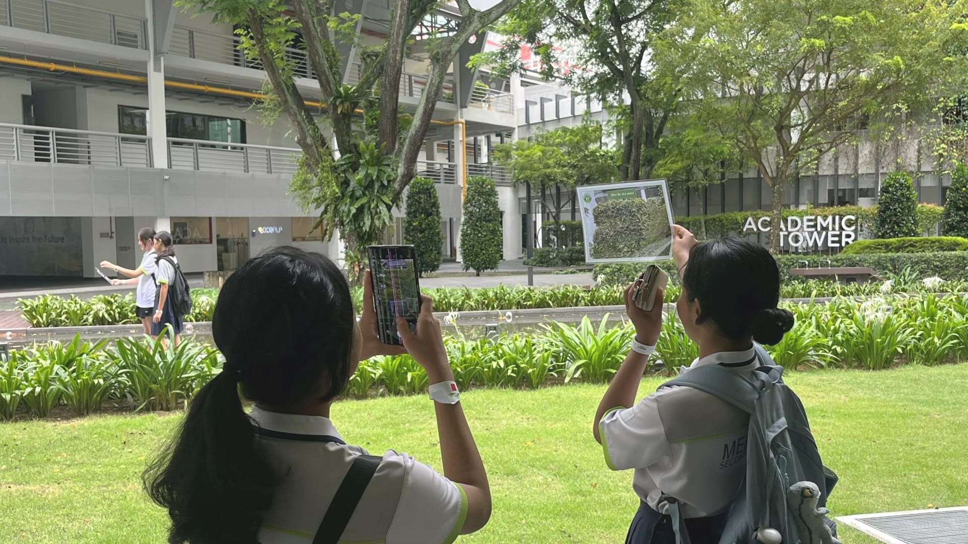 Two students outdoors using mobile devices to capture images of a campus environment, likely as part of a learning activity or workshop.
