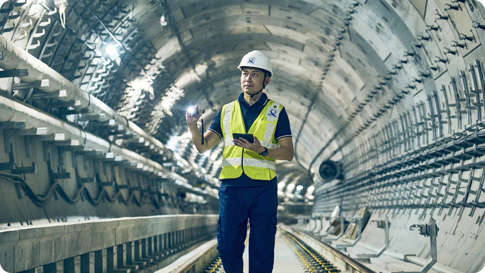 A person in a yellow safety vest and white helmet holds a flashlight and tablet inside a tunnel with illuminated walls and tracks.