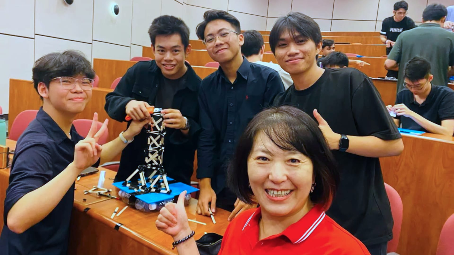 A group of students and a teacher in a lecture hall smiling and posing with a small engineering model on a desk, showing teamwork and hands-on learning.