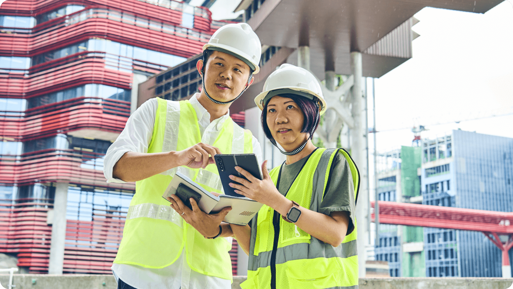 Two people wearing hard hats and yellow vests are looking at a tablet outside near construction buildings.