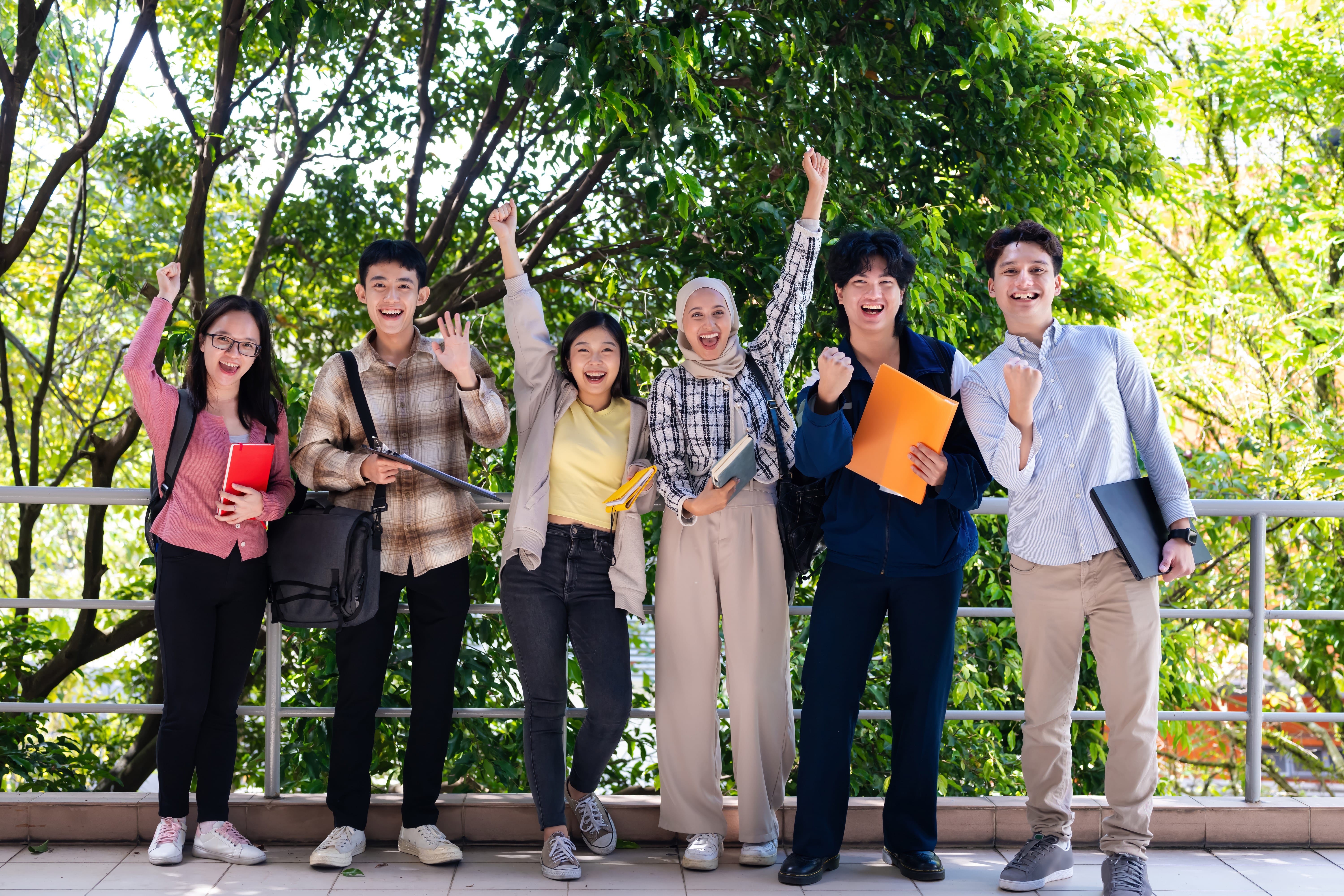 A diverse group of six young students standing outdoors under trees, smiling and cheering with their hands raised, holding notebooks, folders, and a laptop, conveying excitement and teamwork.