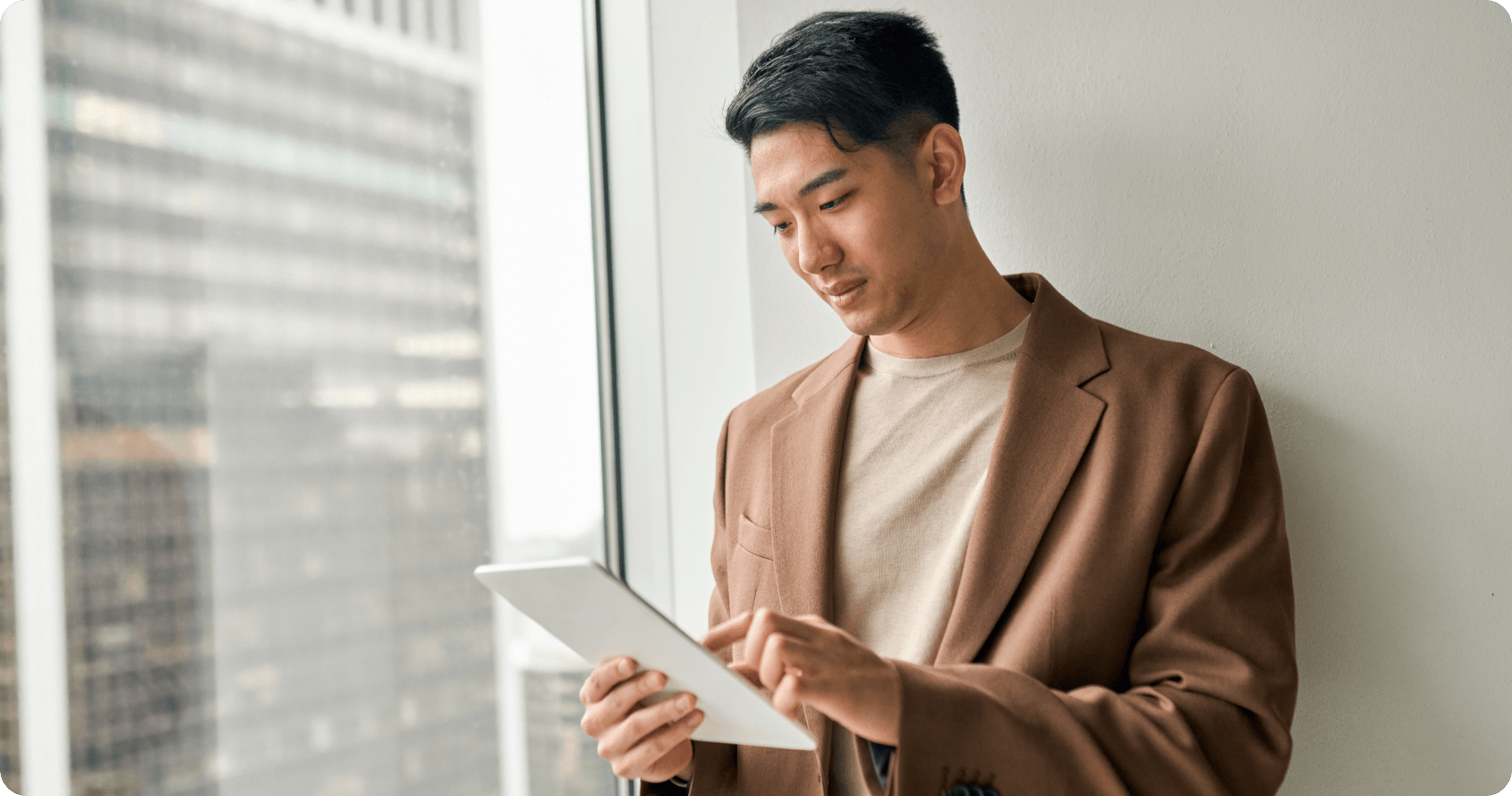 A Built Environment professional in a brown blazer and beige shirt holds a digital tablet while standing by a large window with Singapore's urban skyline visible in the background, representing the continuous learning and career development opportunities available to existing sector professionals. This scene illustrates how Built Environment practitioners can leverage technology and ongoing education to advance their expertise and explore diverse roles within Singapore's dynamic construction industry.