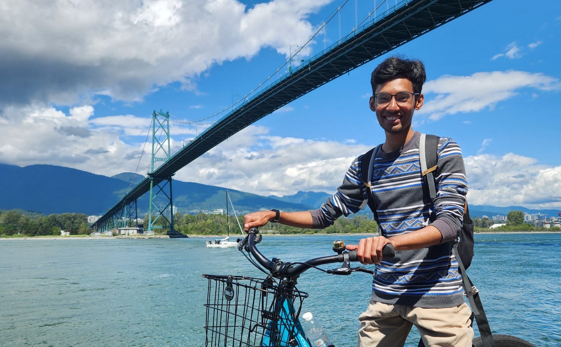 Student posing with a bicycle beside a river under a suspension bridge, with blue skies and mountains in the background, suggesting travel or outdoor exploration.