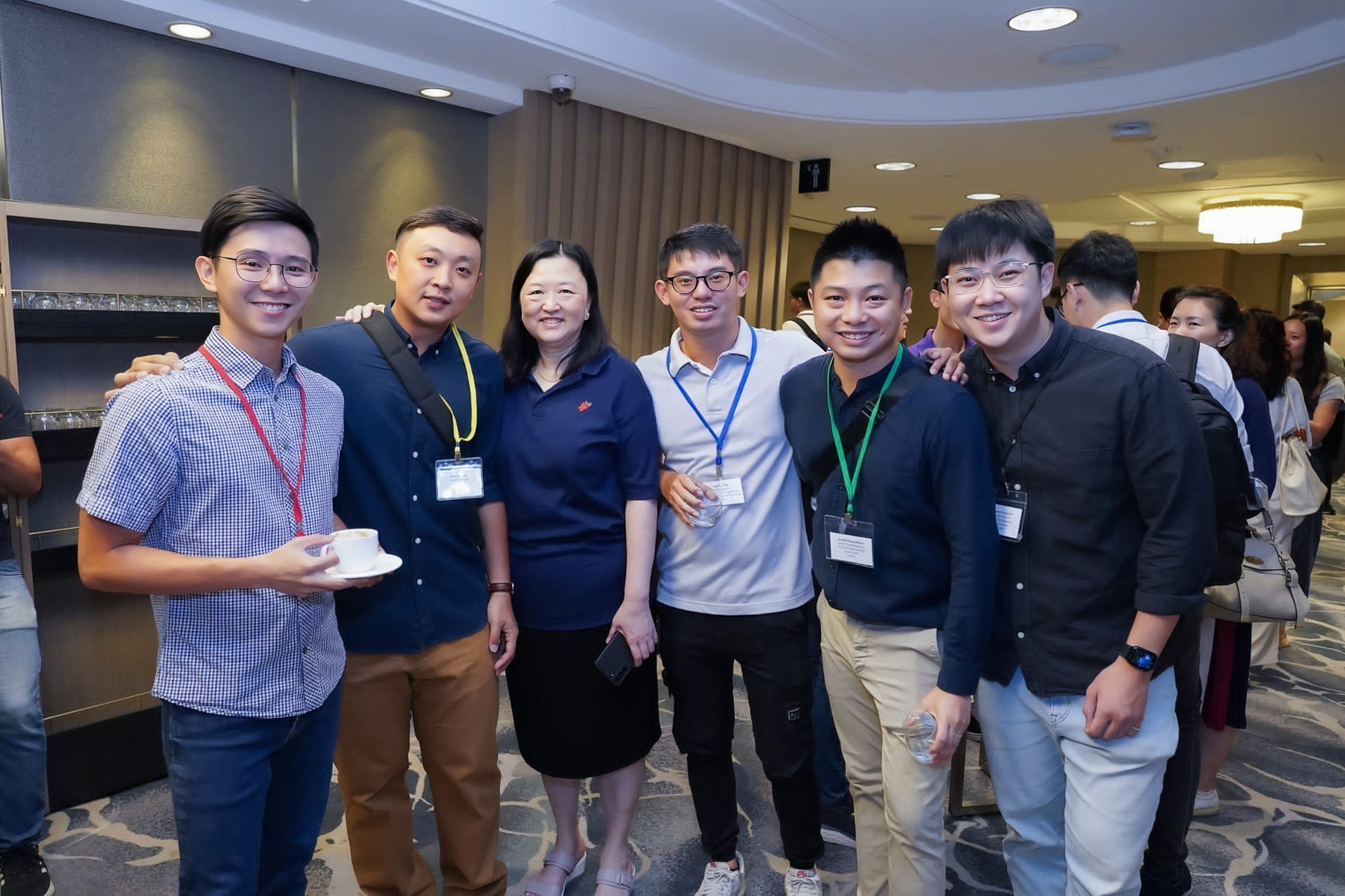 Six professionals with name tags smiling together at a networking event, standing in a conference venue with drinks in hand and other attendees in the background.