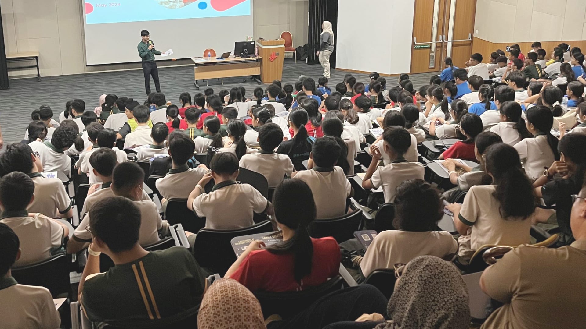 A large group of students seated in lecture theatre attending a presentation, with a speaker at the front engaging the audience.