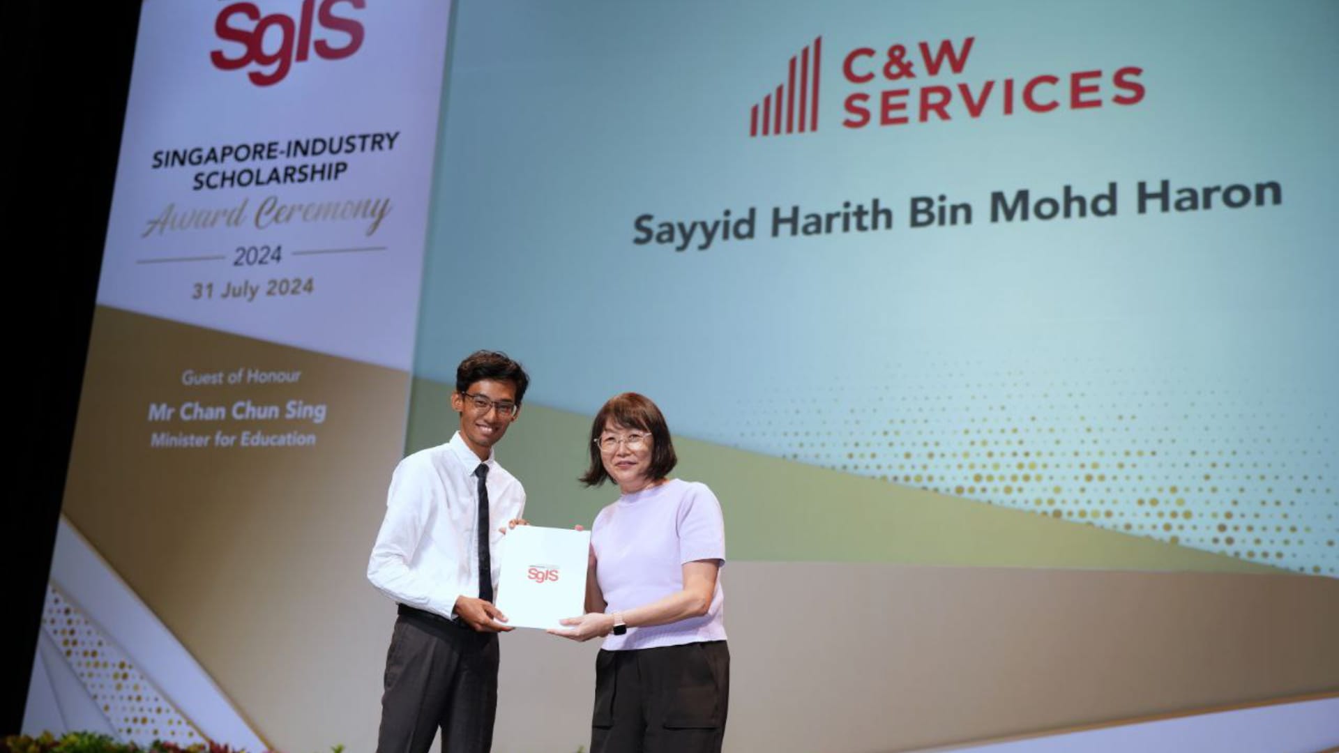A young man receiving a Built Environment scholarship certificate on stage from a presenter, with a backdrop displaying his name and organisation.