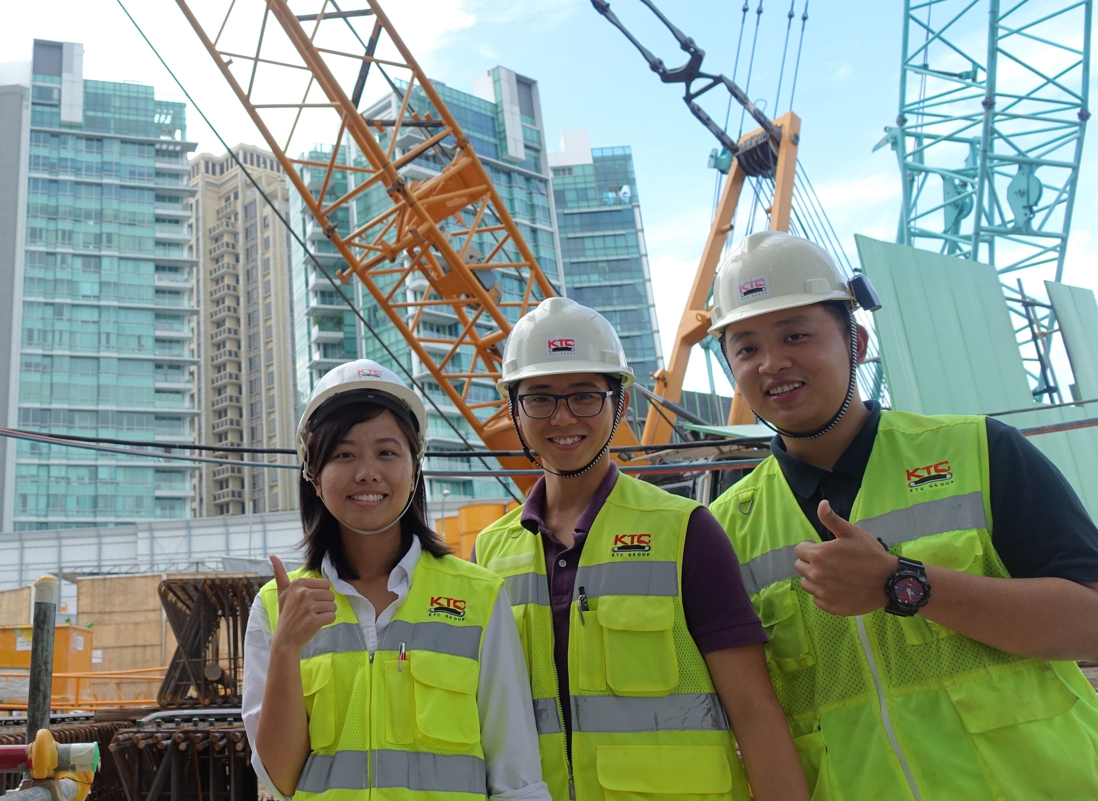 Three construction workers wearing white safety helmets and bright yellow reflective vests stand at a construction site, smiling and giving thumbs up. Behind them are cranes and high-rise buildings under construction.