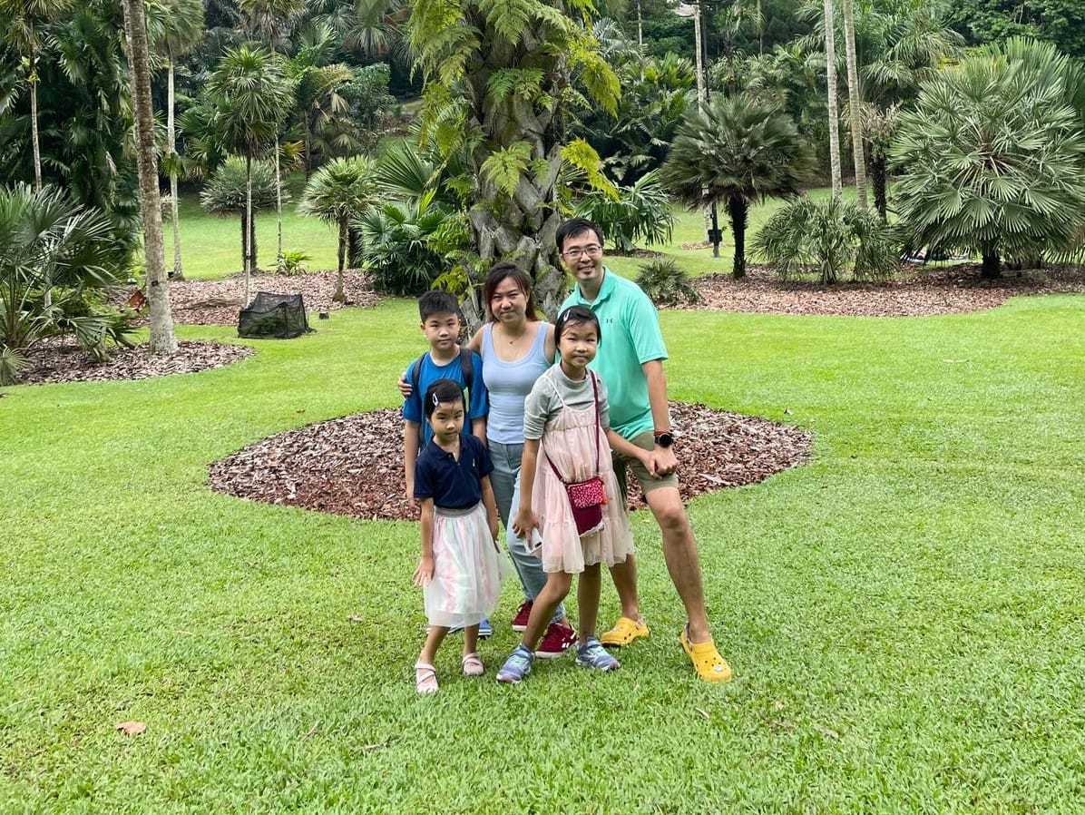 A group of five people standing on green grass with trees and bushes in the background at a park.