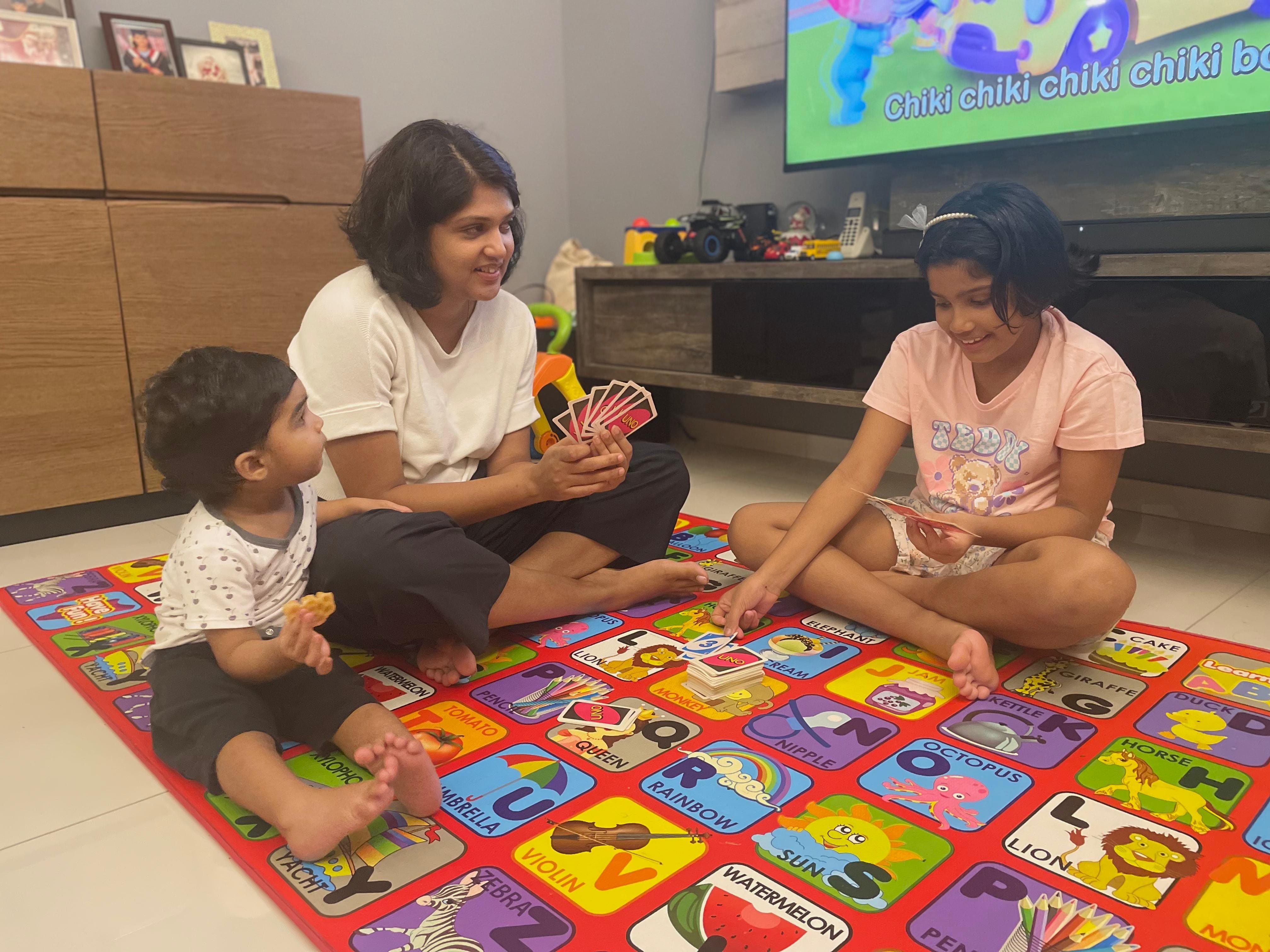 Three people sit on a colorful alphabet mat, holding cards with toys and a TV screen in the background.