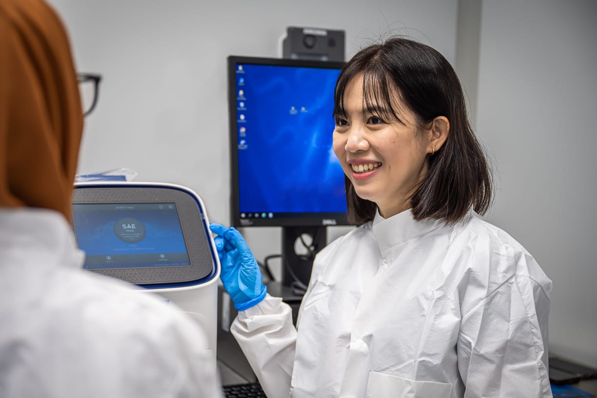 Two people in lab coats and gloves work with a scientific machine, with a monitor displaying a blue screen in the background.
