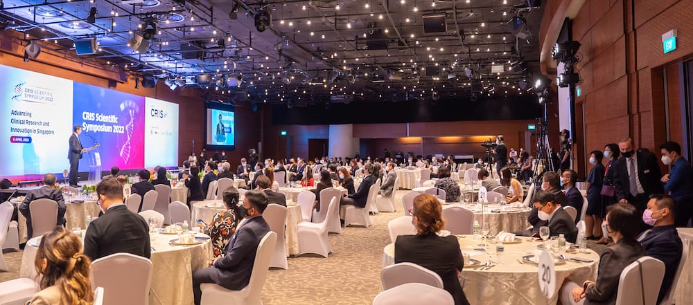 A conference room filled with people seated at round tables, a speaker on stage, and screens displaying CRIS Scientific Symposium 2022.