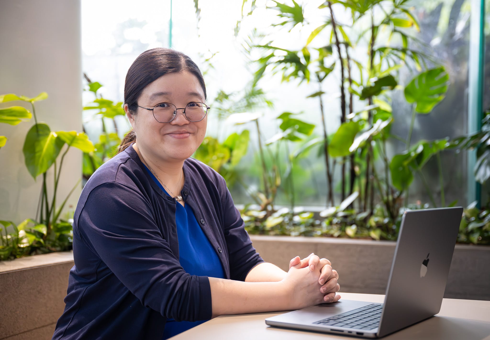 A person wearing a blue blouse and navy cardigan is sitting at a table with a laptop, surrounded by plants.