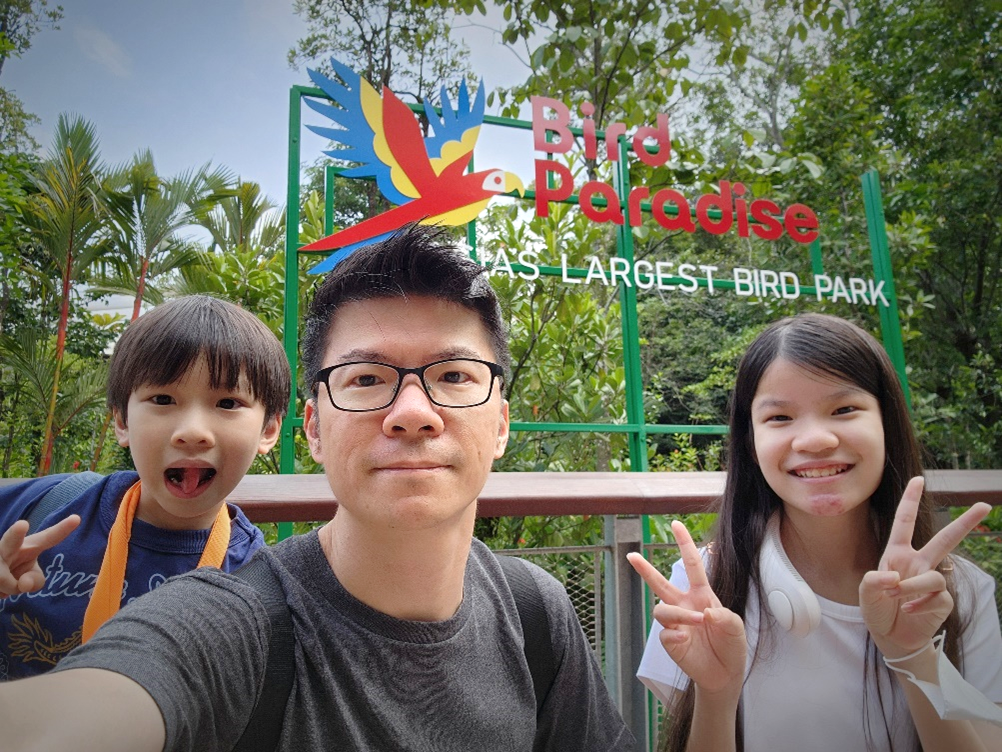 Three people make peace signs in front of a colorful "Bird Paradise" sign surrounded by trees.