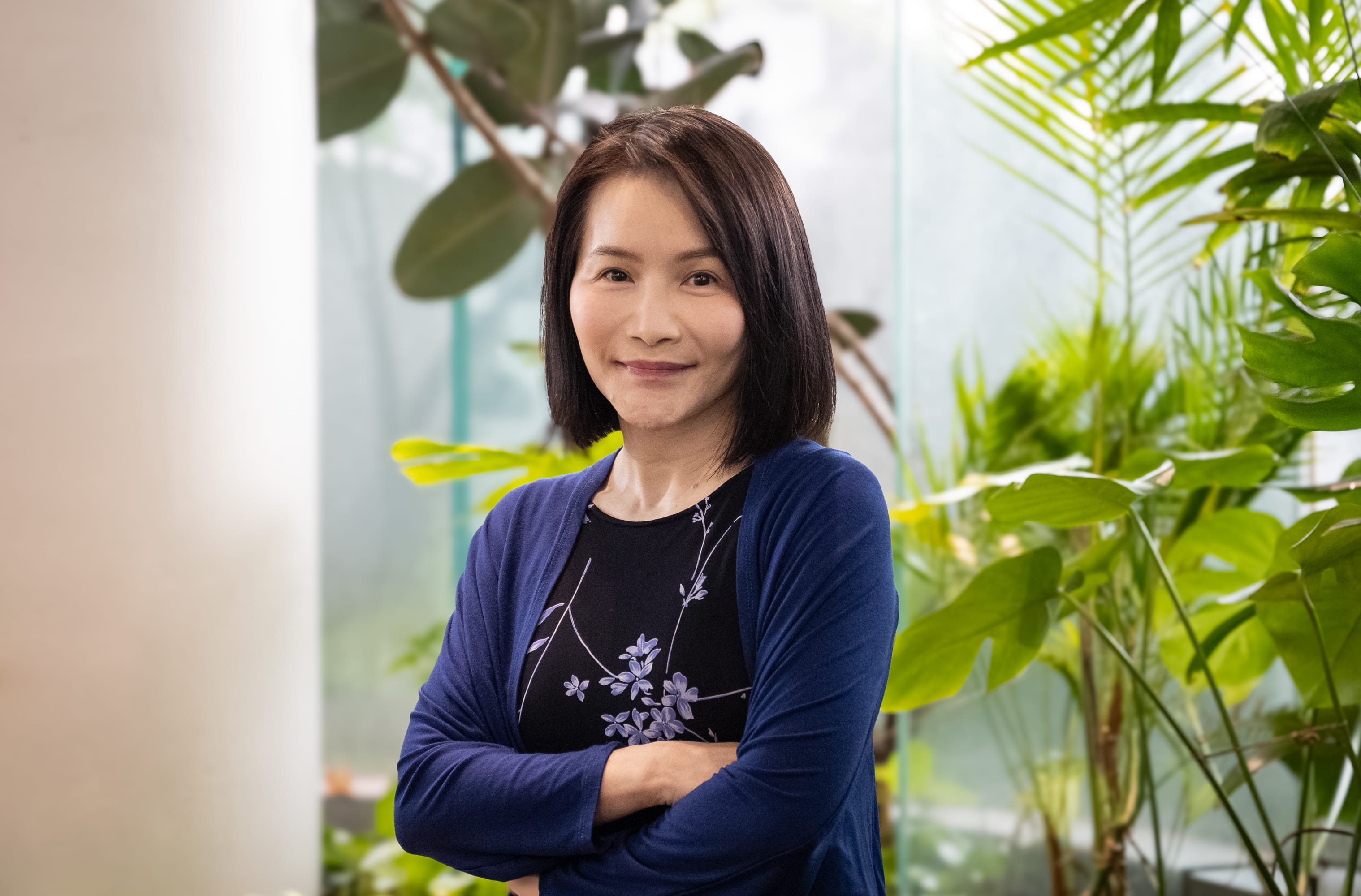 Person standing with arms crossed, wearing blue cardigan over black dress with floral pattern, surrounded by leafy plants.