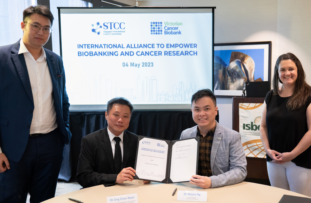 Two men in suits are seated at a table holding a signed document, while two people stand behind them; a presentation screen shows biobanking details.