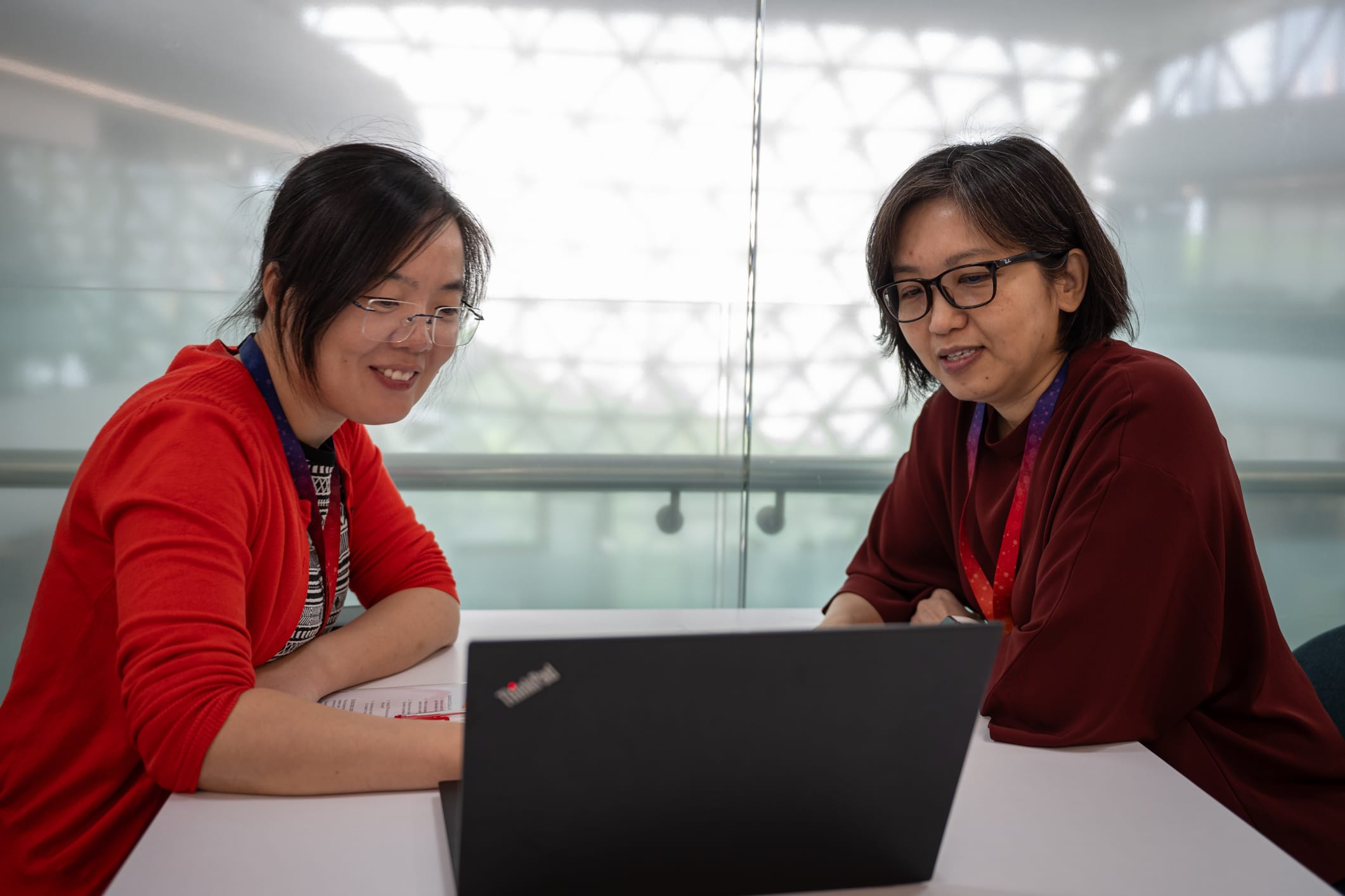 Two people are sitting at a table with a laptop between them, one wearing a red cardigan, the other a red shawl.