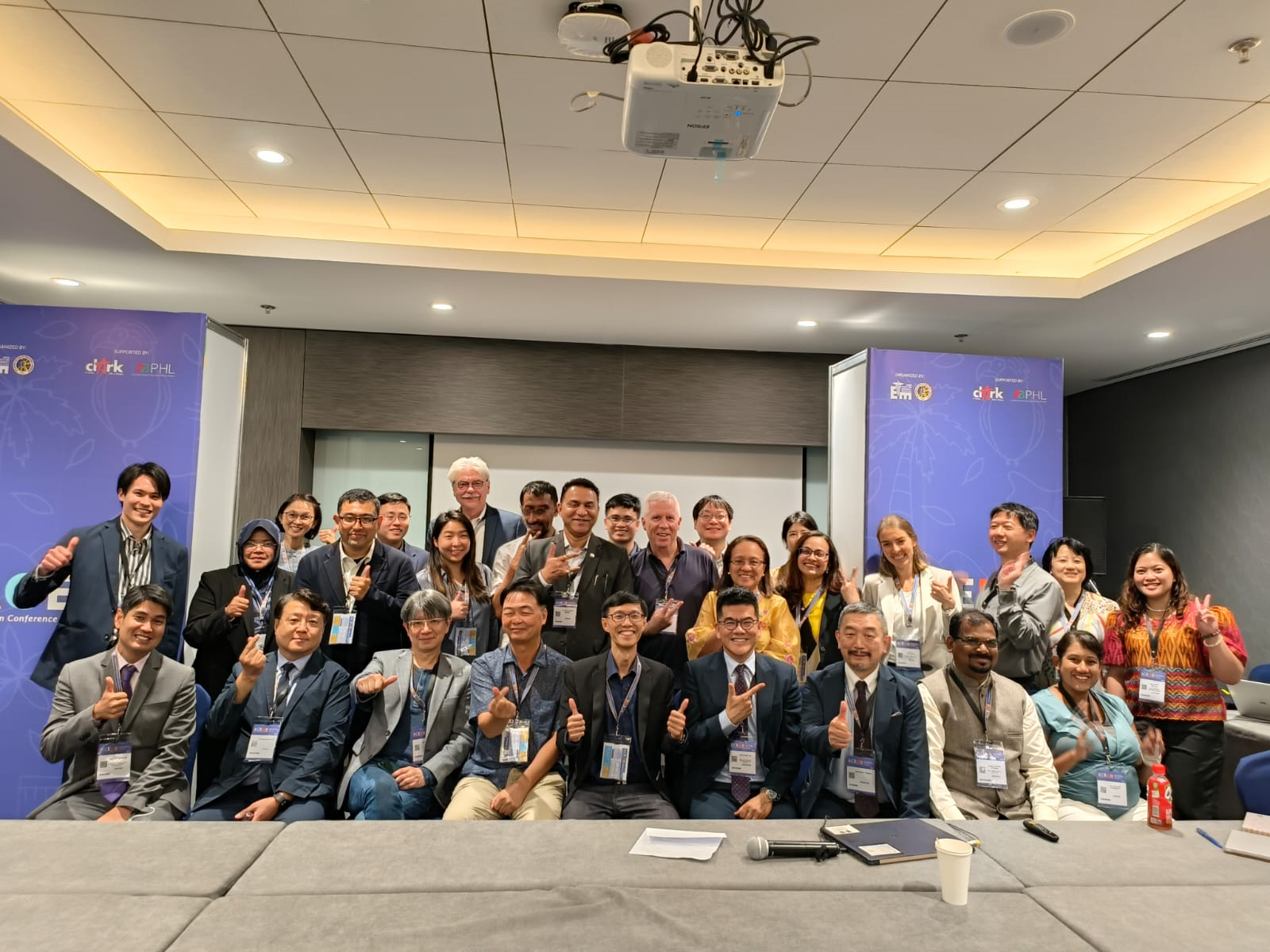 A group of roughly 20 people posed together in a conference room, several making thumbs-up gestures, with presentation equipment above.