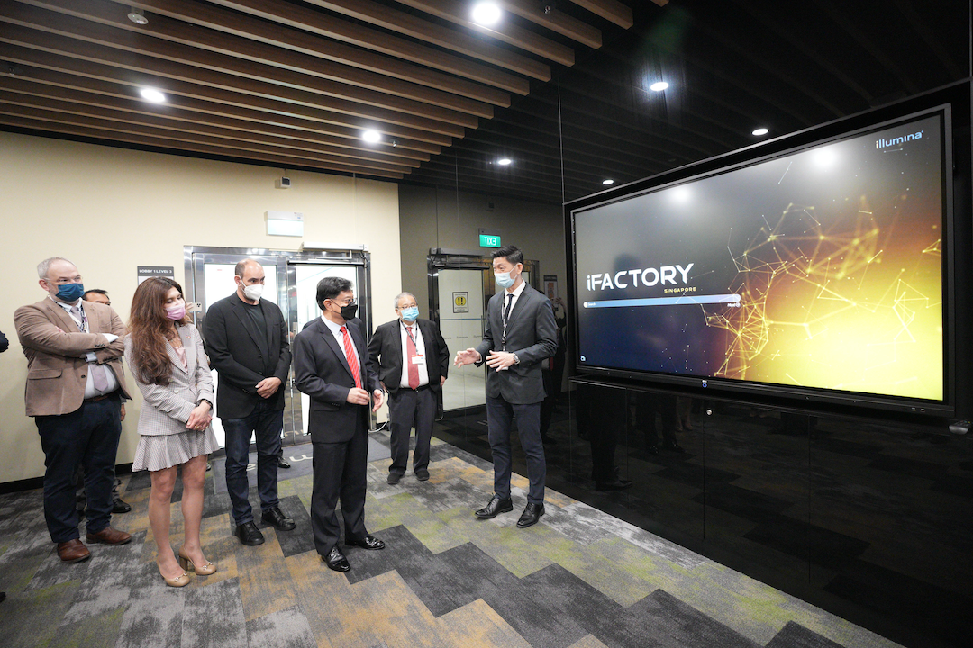 Seven people stand in front of a large screen displaying "iFACTORY SINGAPORE" in an office setting.
