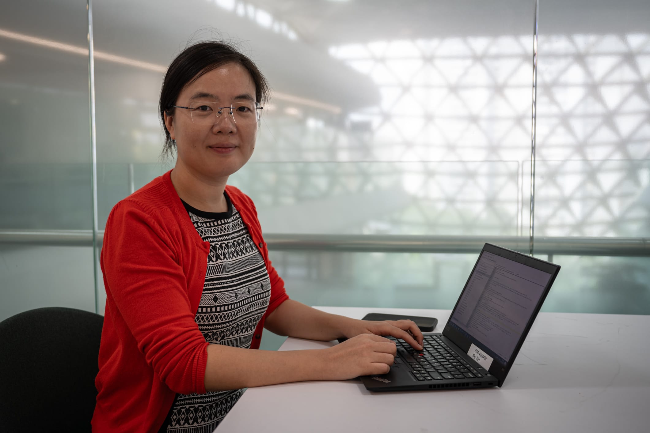 A person in a red cardigan and patterned shirt types on a laptop at a table in a modern, bright office setting.