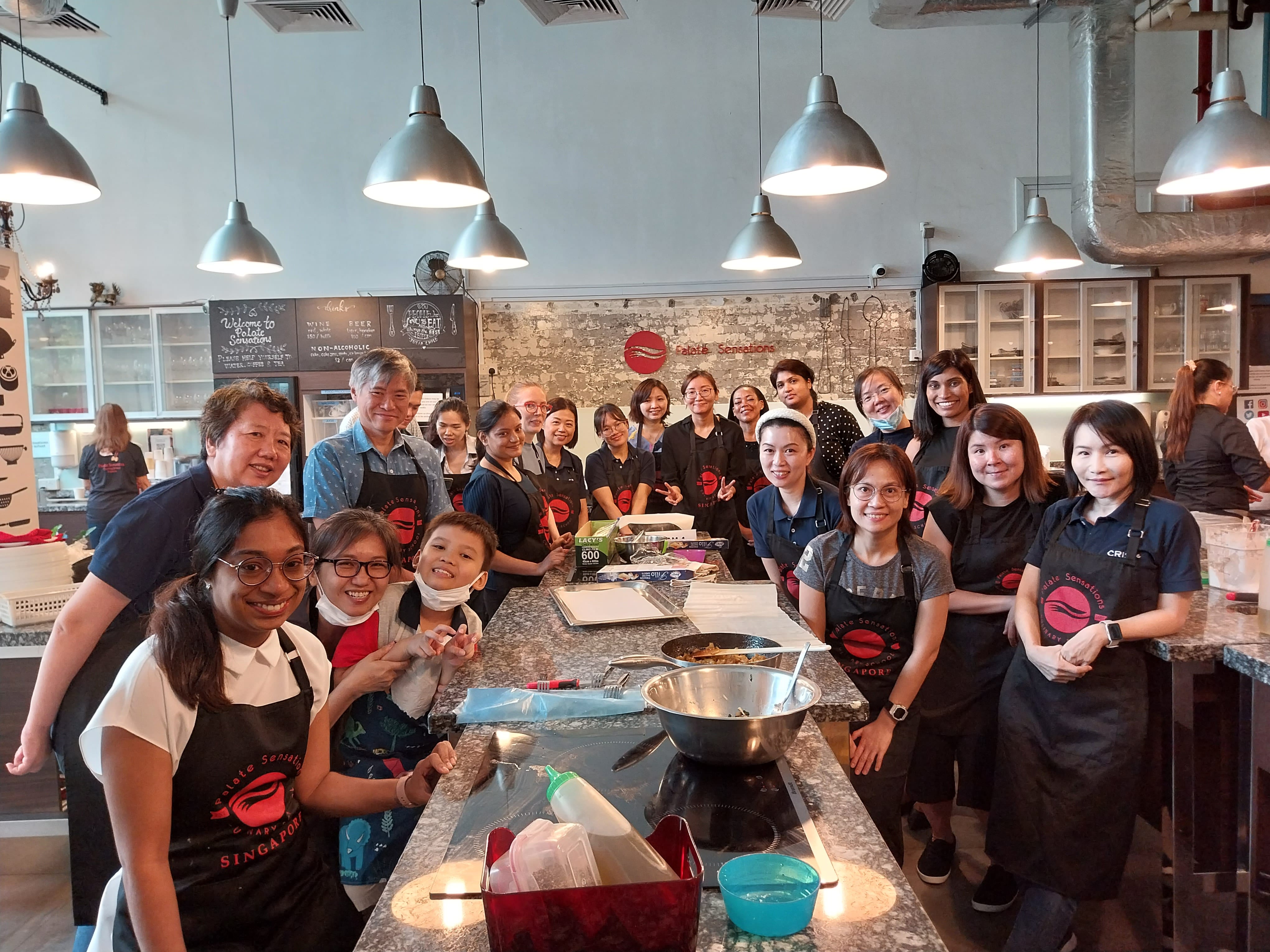 A group of people wearing aprons gather around a kitchen counter with bowls and utensils, under industrial hanging lights.