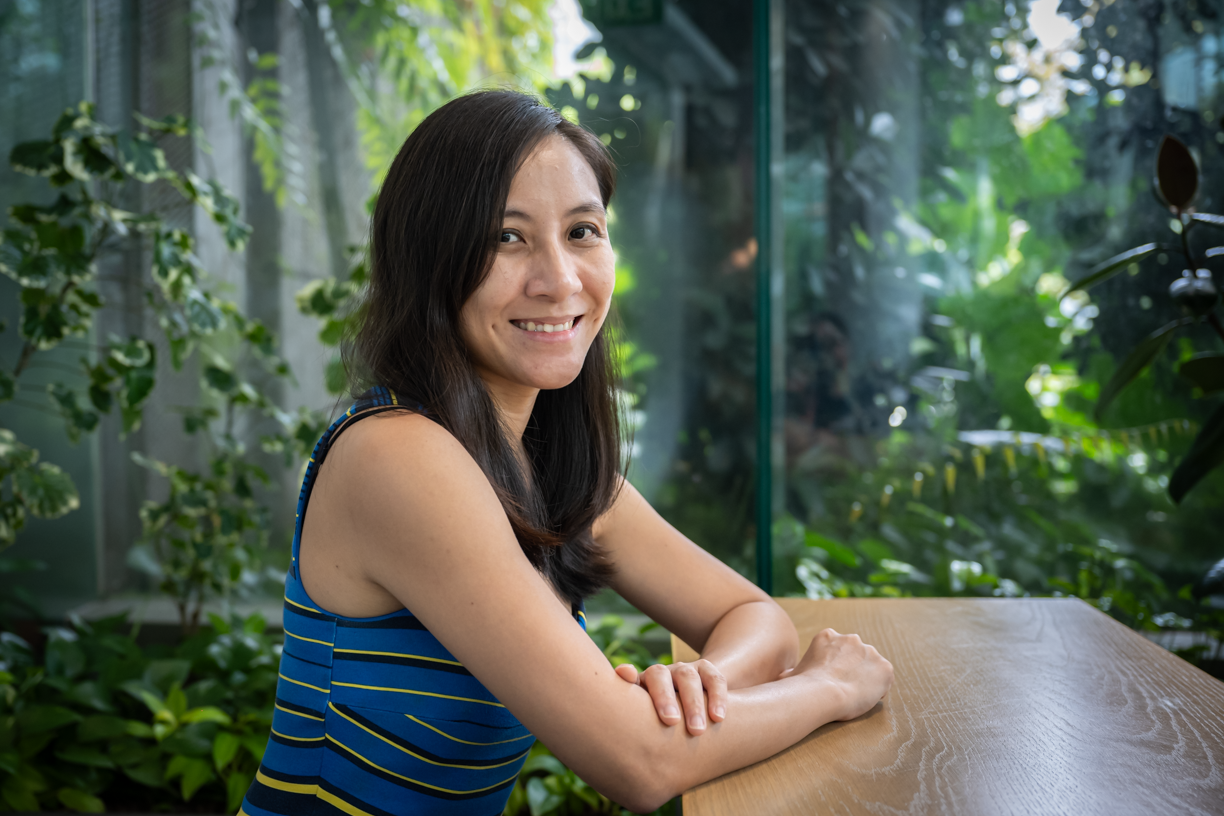 A woman with long dark hair sits at a wooden table in a lush indoor garden.