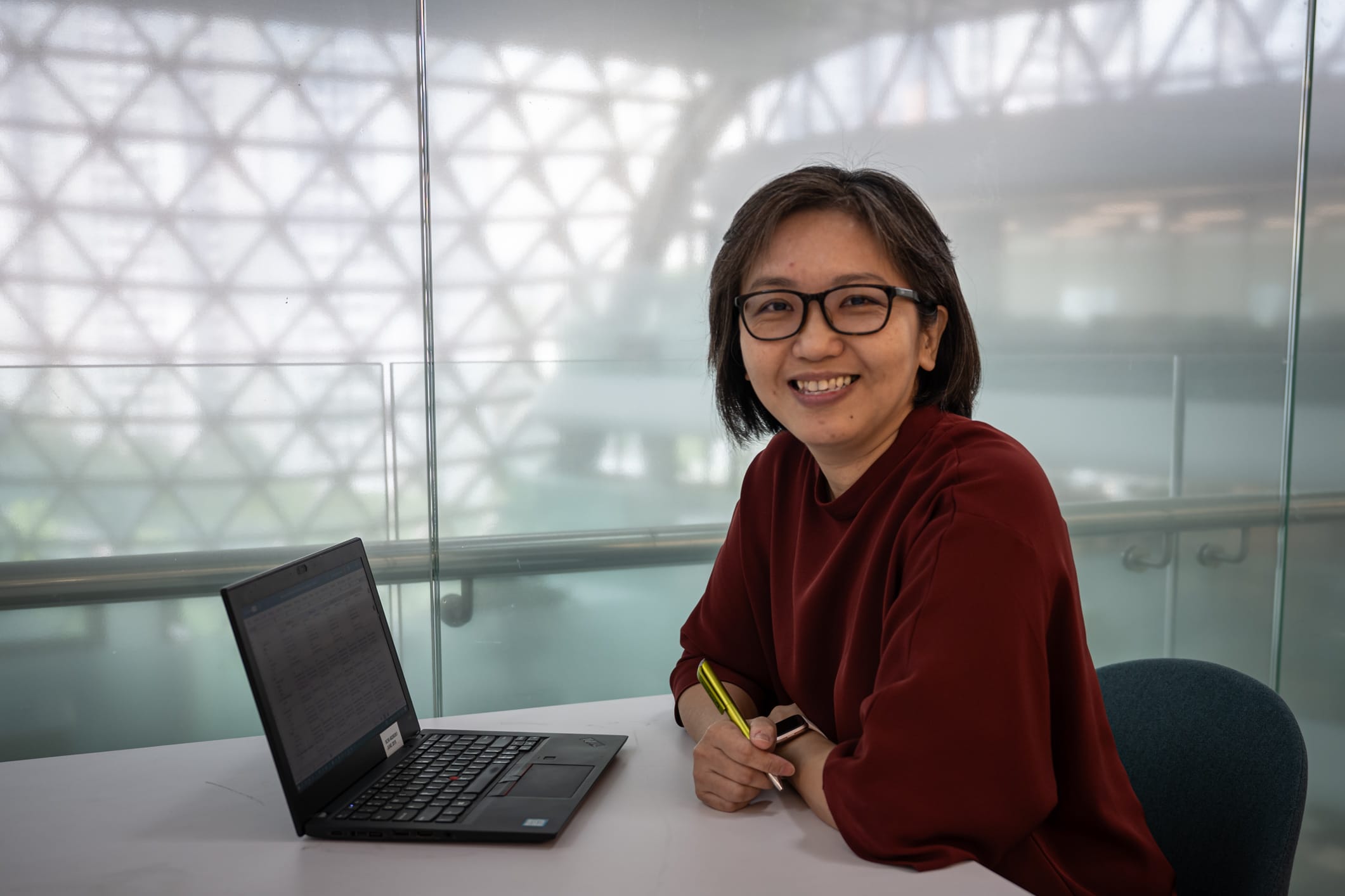 A person wearing a red shirt is sitting at a desk with a laptop and holding a yellow pen.