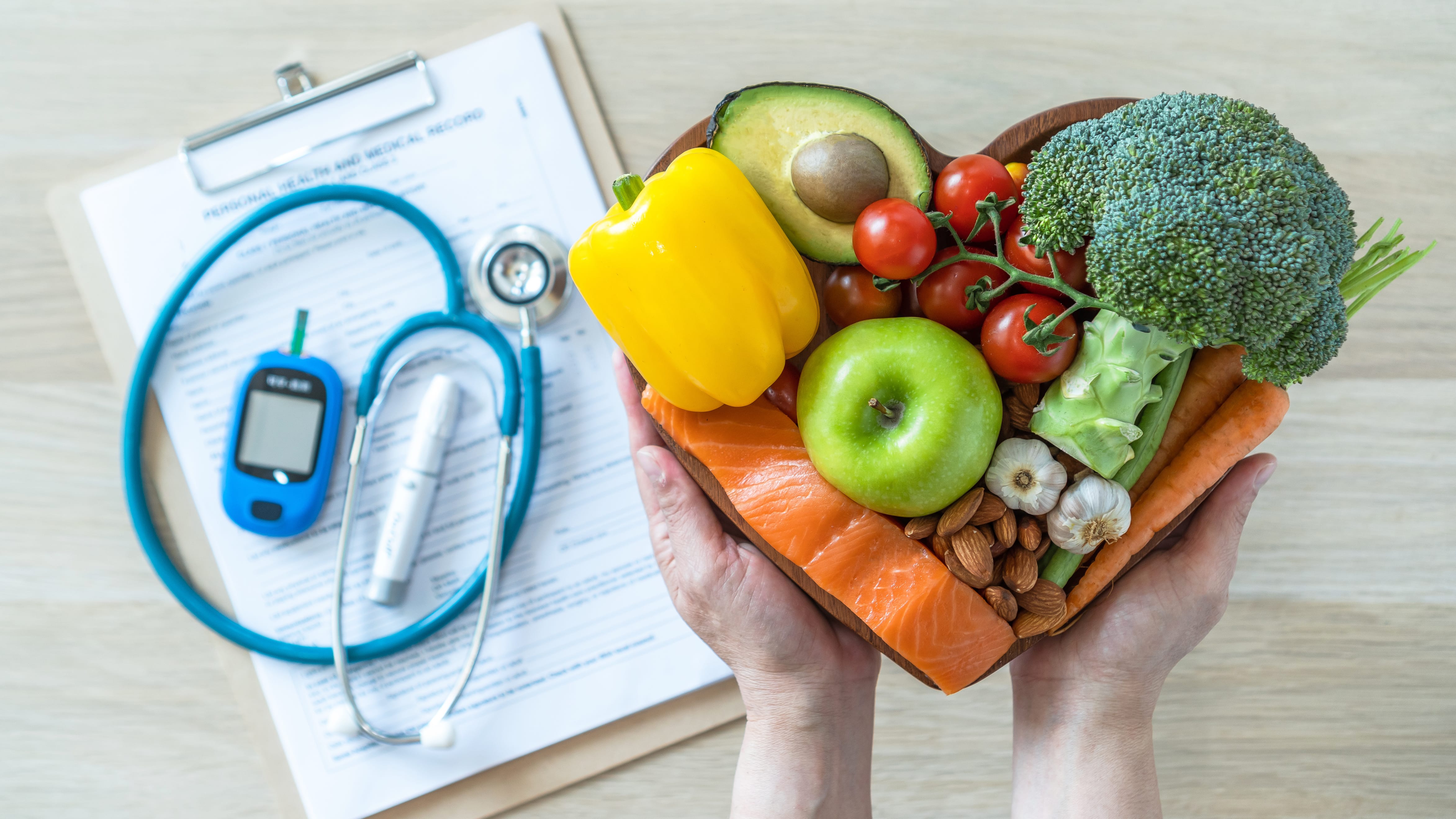 Hands holding a bowl of assorted vegetables, fruits, nuts, and salmon, with medical equipment and a clipboard in the background.