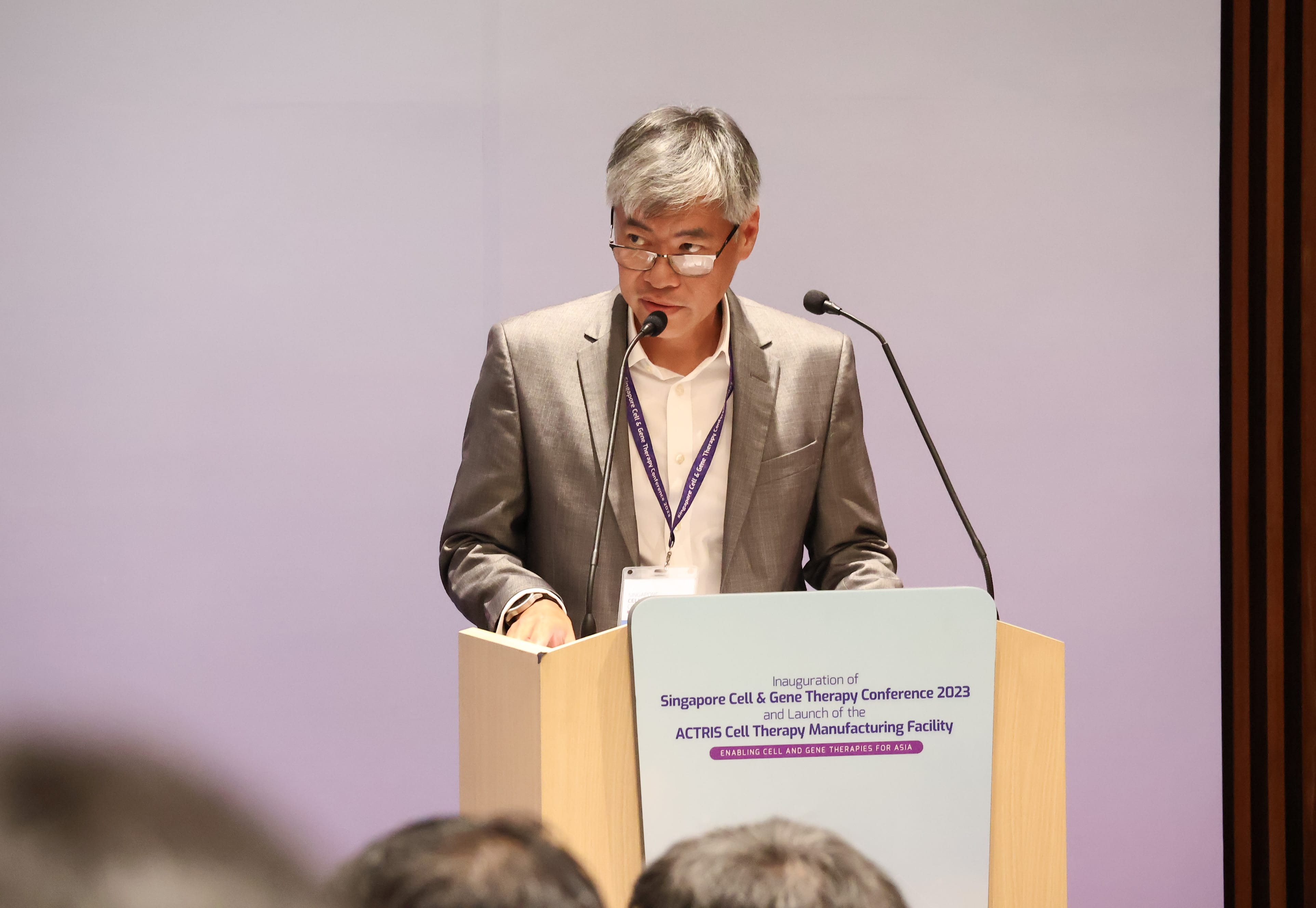 A person wearing a suit and conference badge stands behind a podium with a microphone and event information.