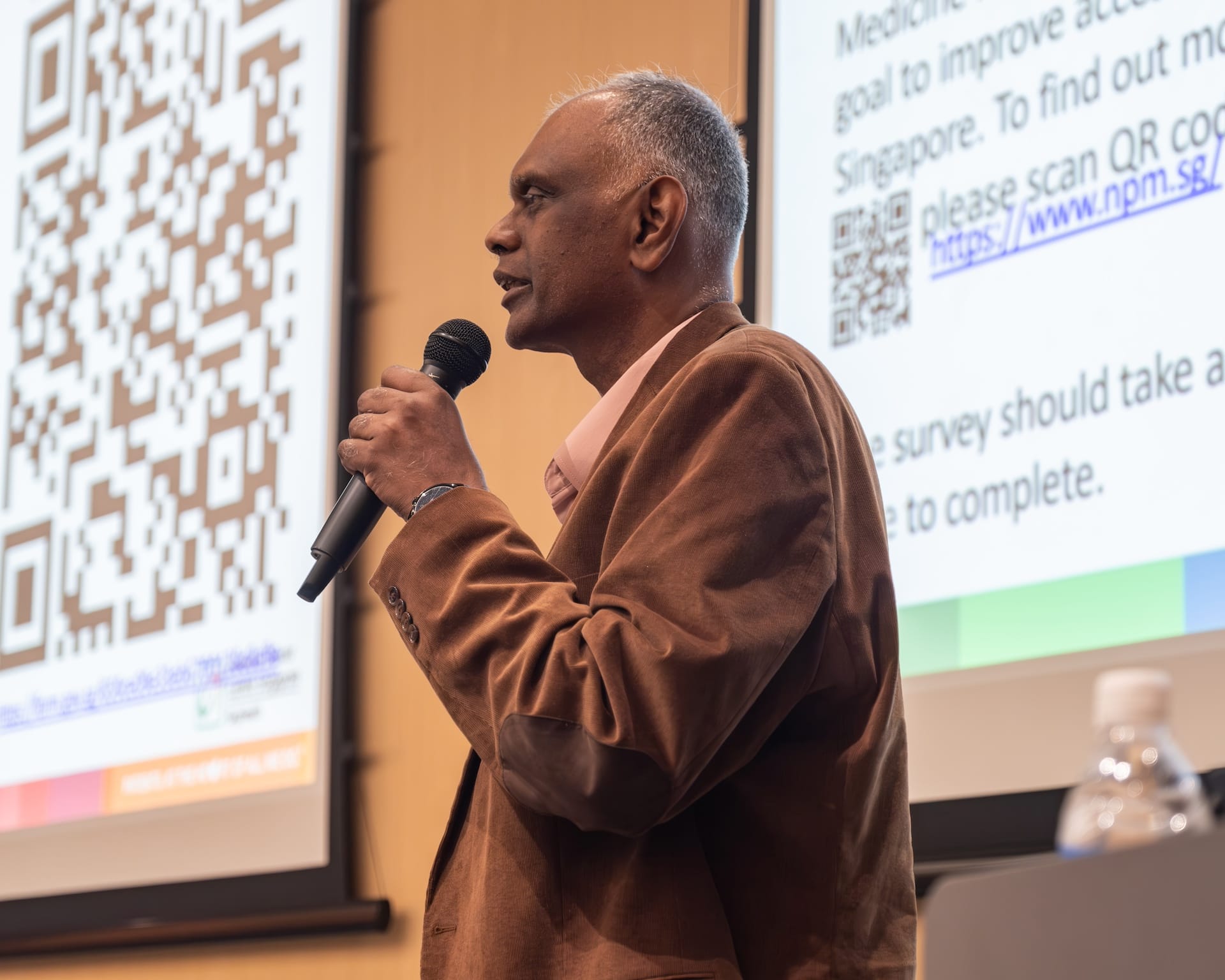 A person in a brown blazer holds a microphone near screens displaying QR codes and text in a presentation room.