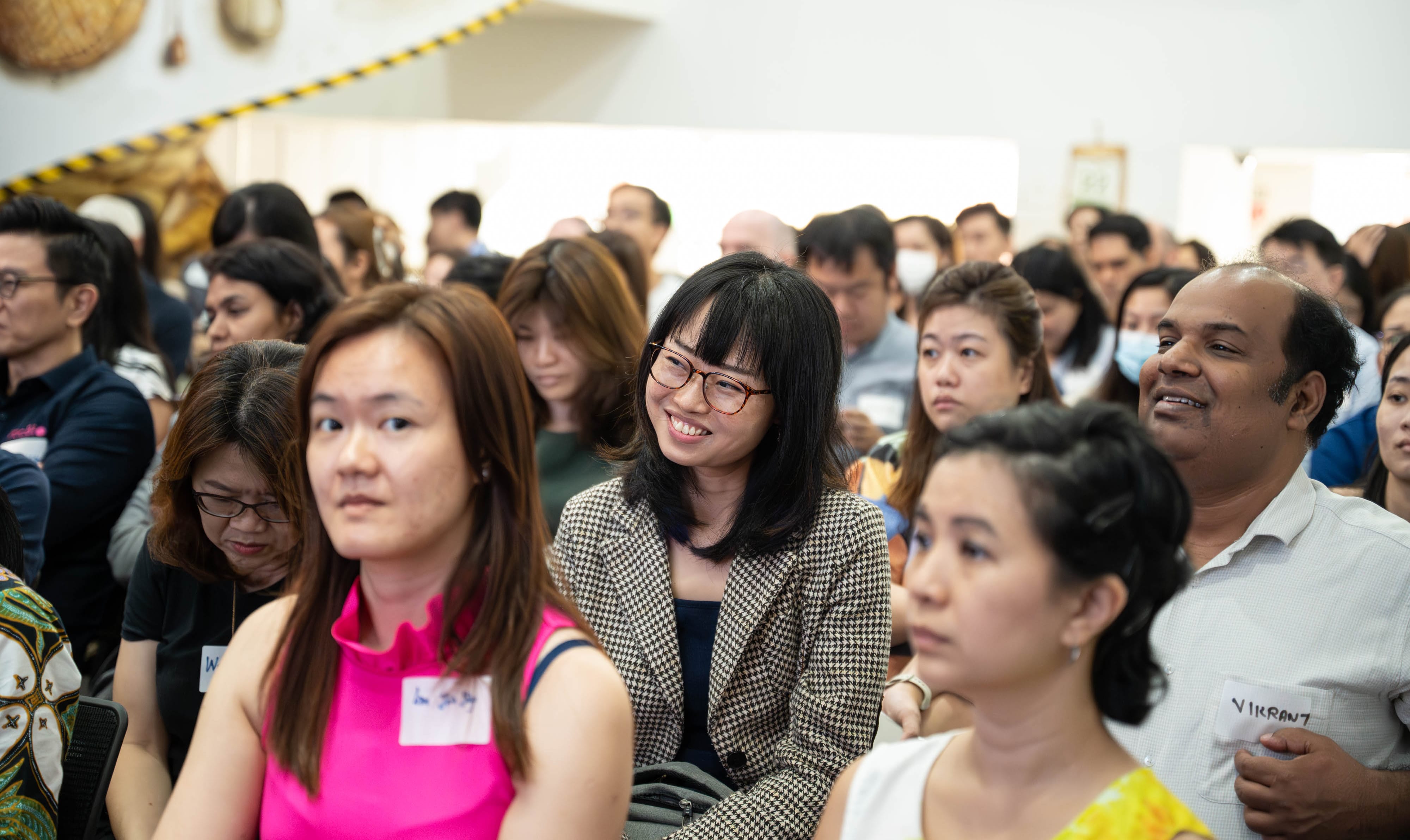 A large group of people are sitting indoors, many wearing name tags, with some visible background decorations.