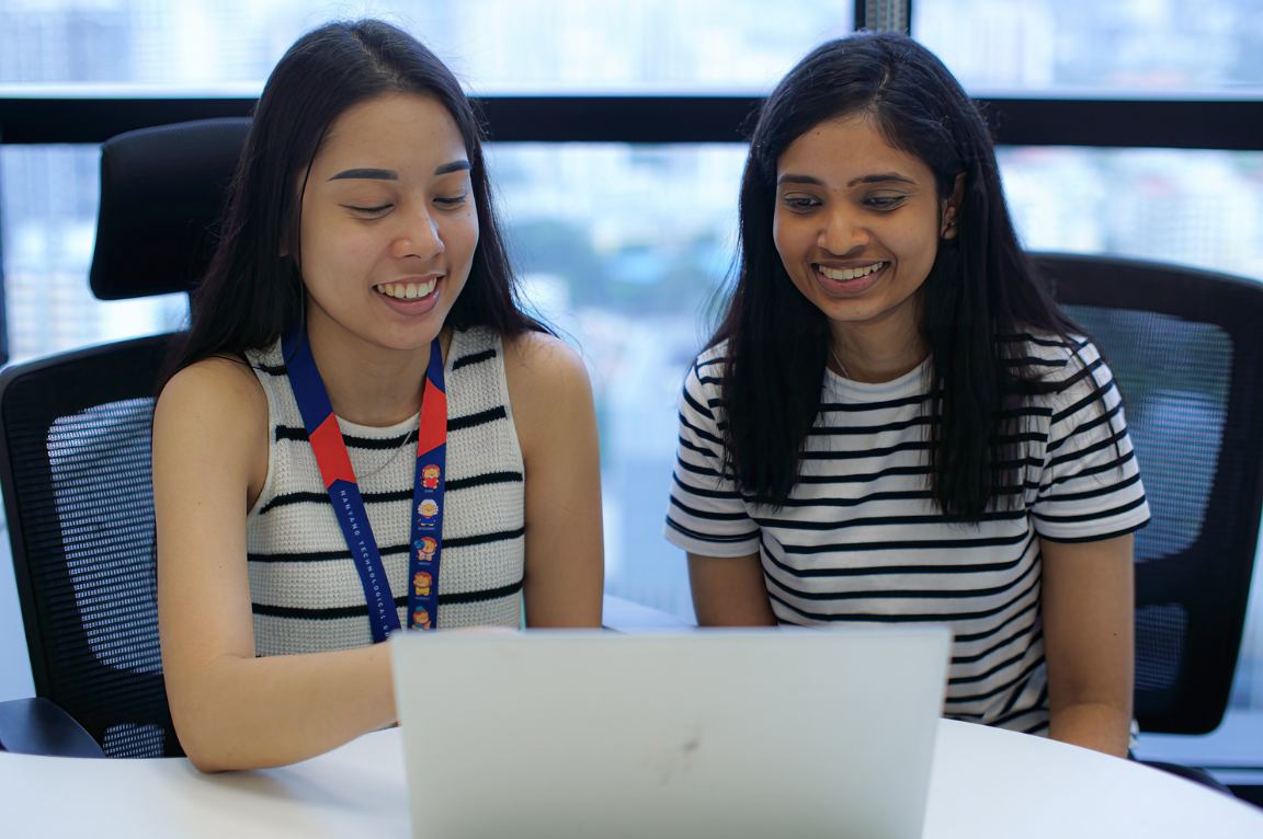 Two women wearing striped tops sit side by side at a table with a laptop in front of them in a bright office.