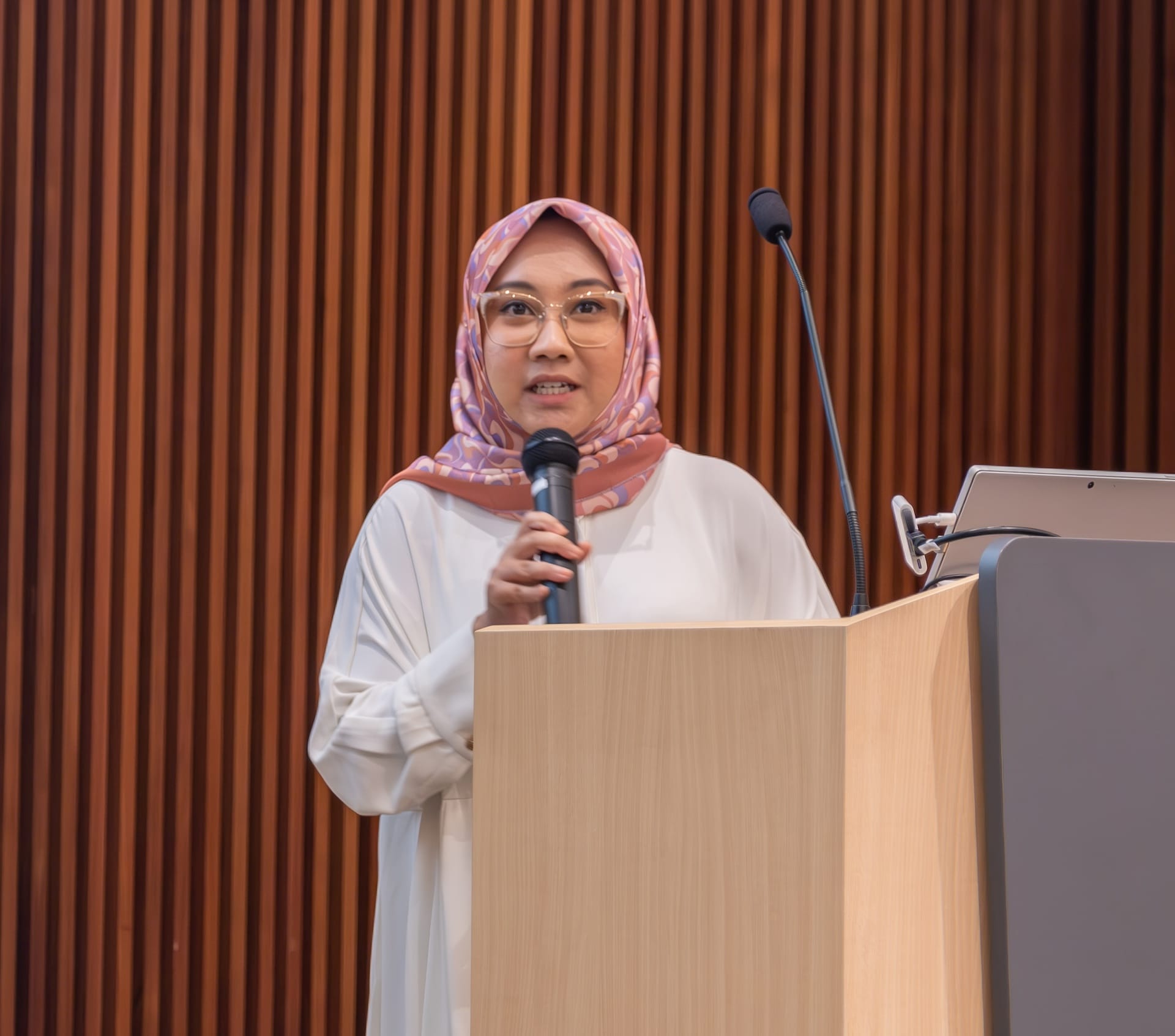 A person wearing a hijab and white clothing stands at a lectern holding a microphone, with a wood-paneled background and a laptop.