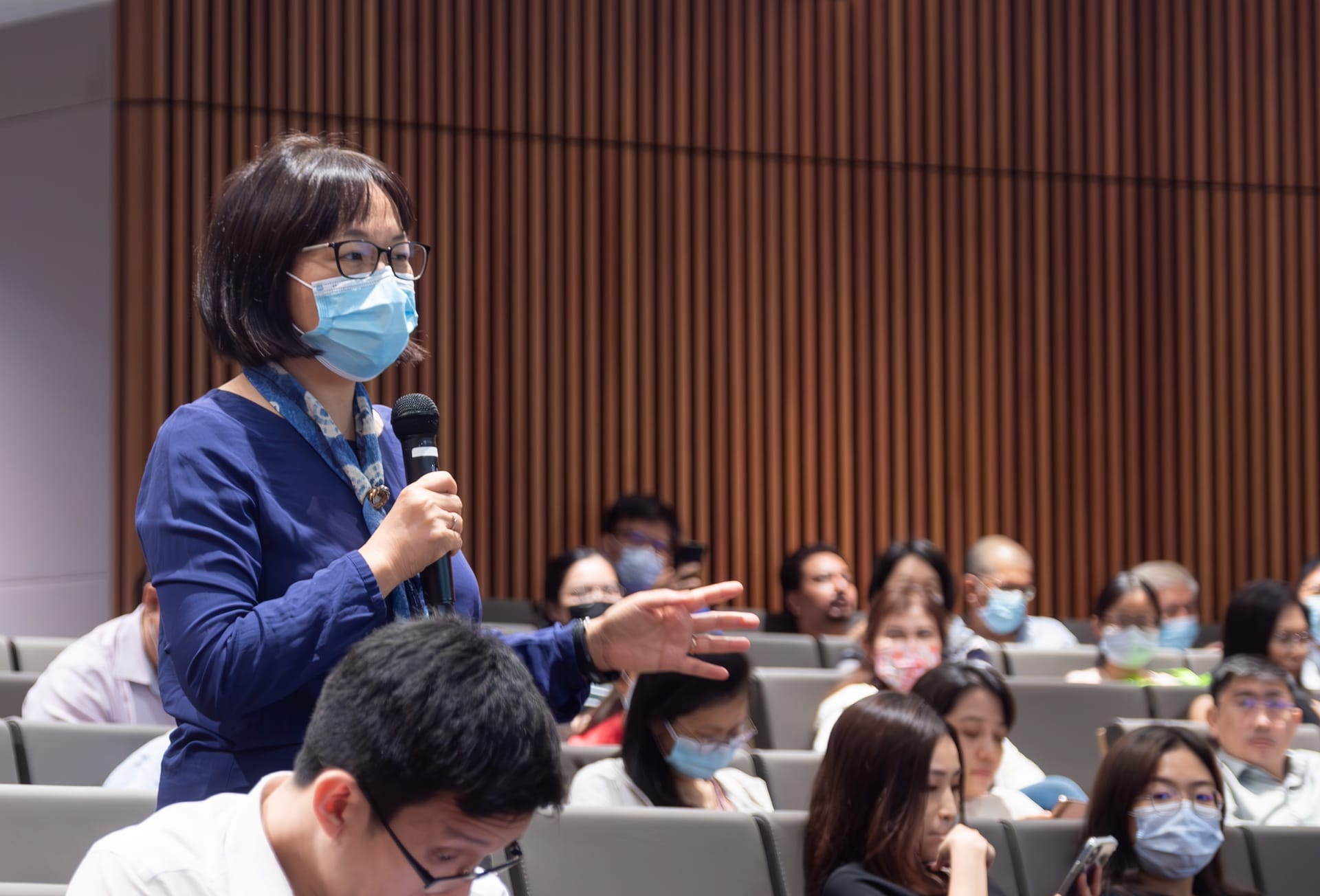 Person in a blue shirt speaking into a microphone to an audience in an auditorium with wooden paneling.