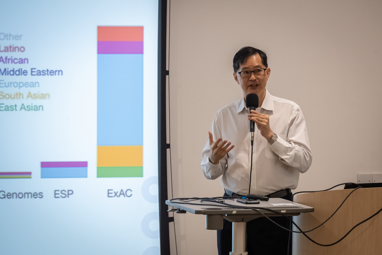 A person standing and speaking at a podium with a microphone, next to a screen displaying a colorful bar chart.