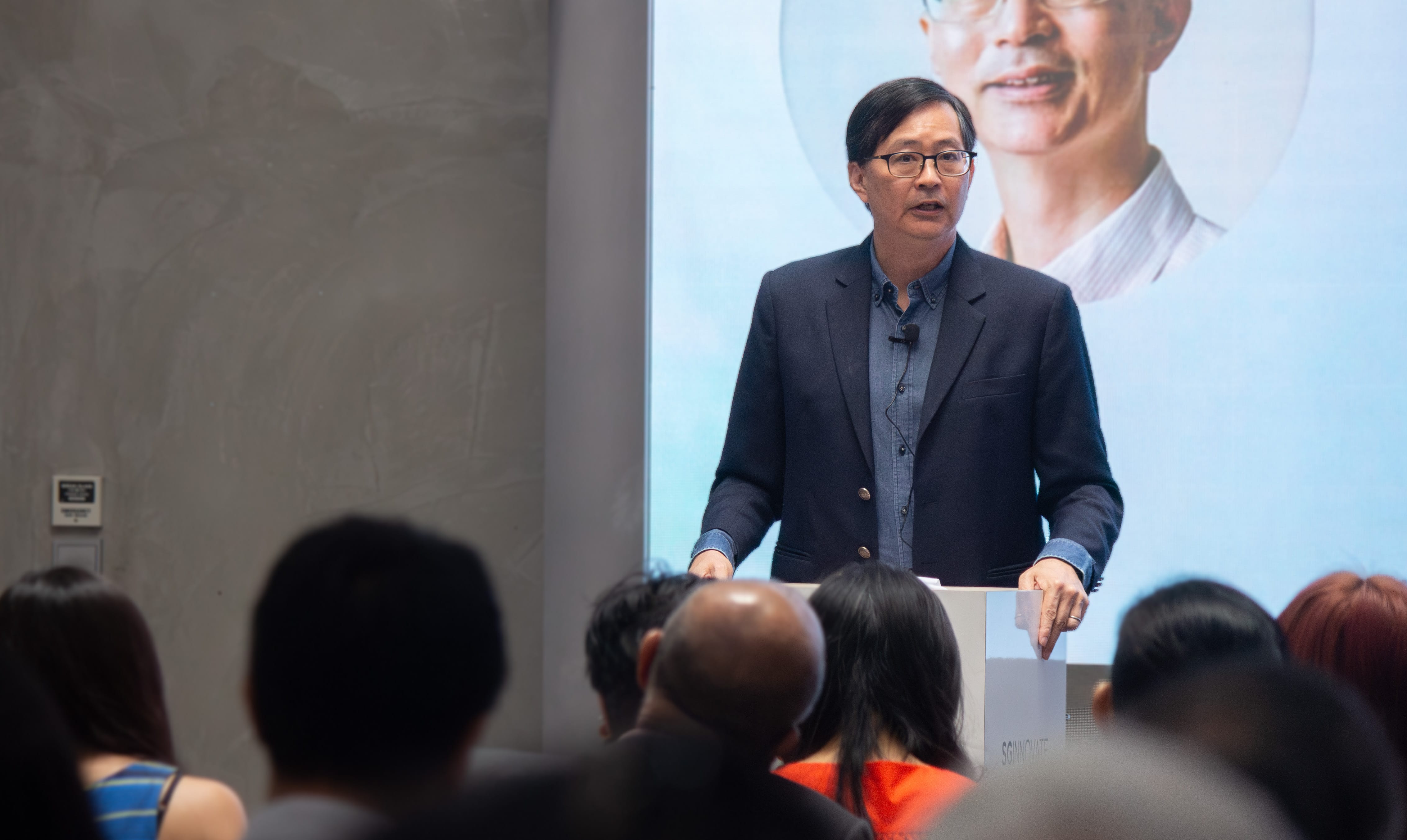A person in a dark blazer and blue shirt is speaking in front of an audience with a large display behind them.
