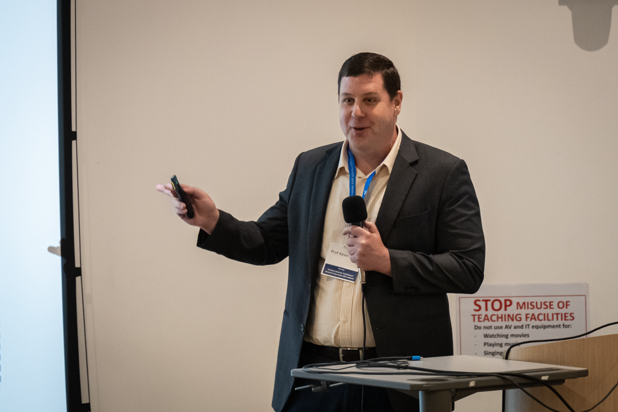 Man in business attire holds a microphone and a remote, standing beside a sign listing prohibited activities.