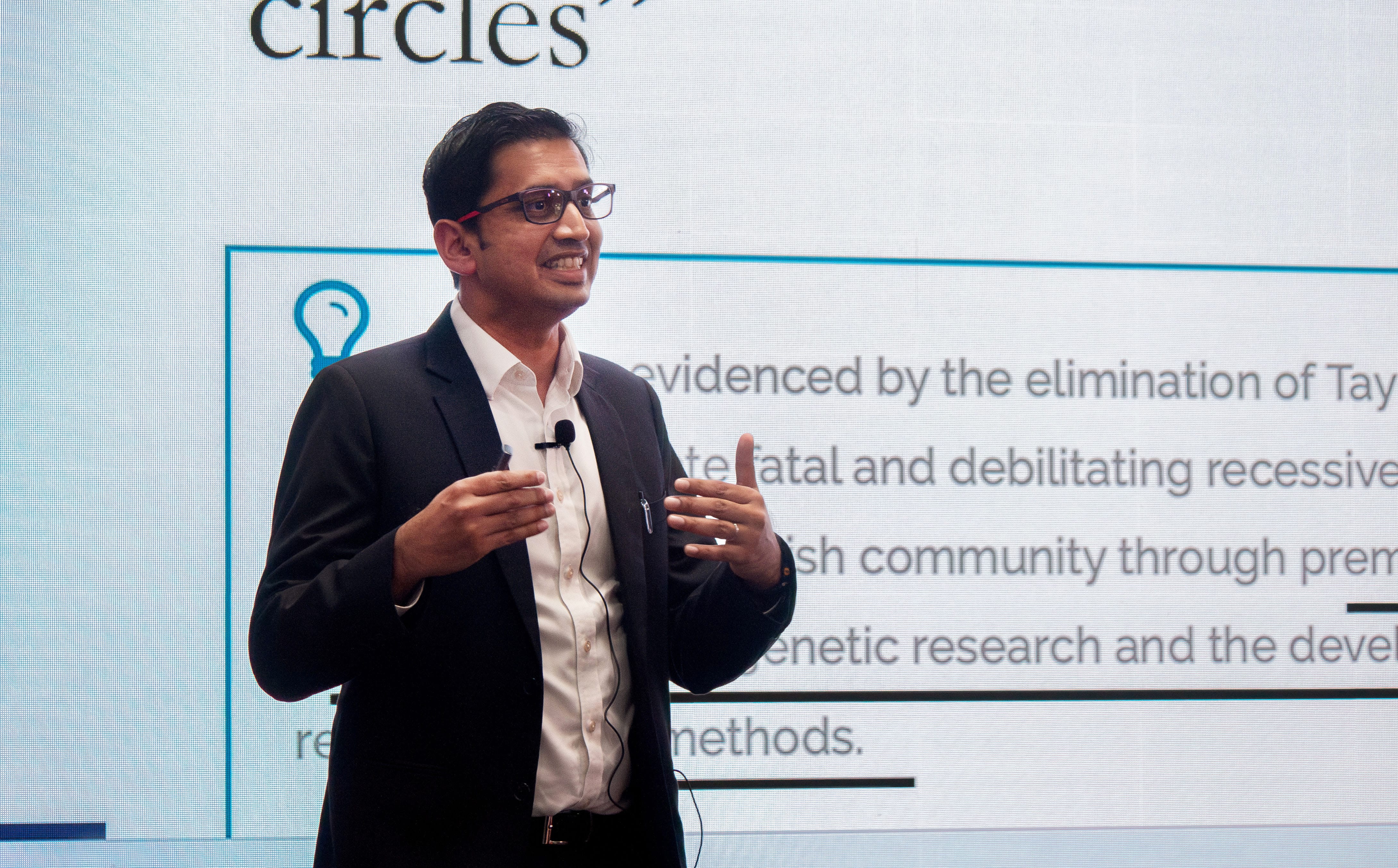 A man in a black suit and white shirt holds a presentation with text projected on the wall behind him.