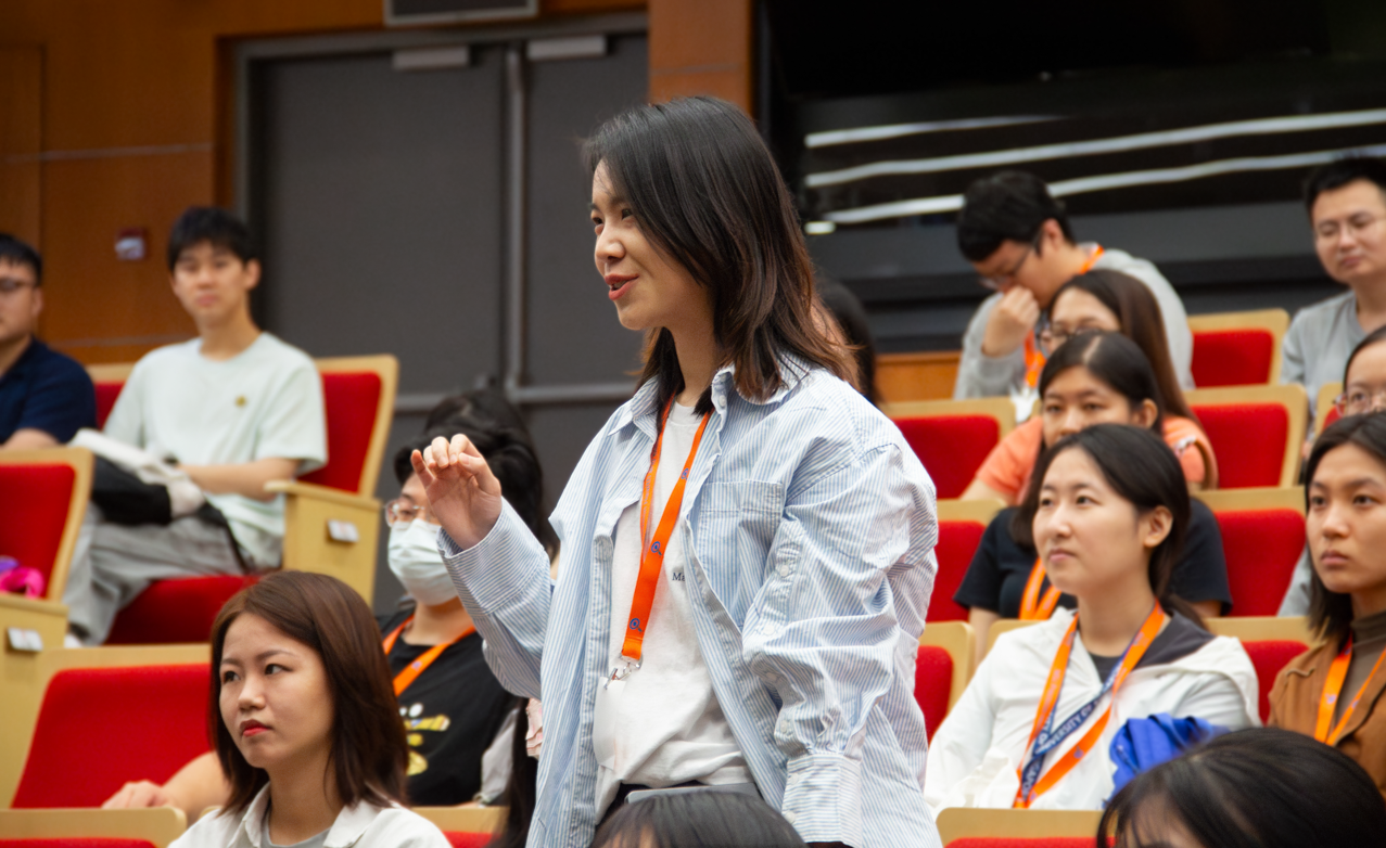 A woman standing and raising her hand in a lecture hall with people seated in red chairs.