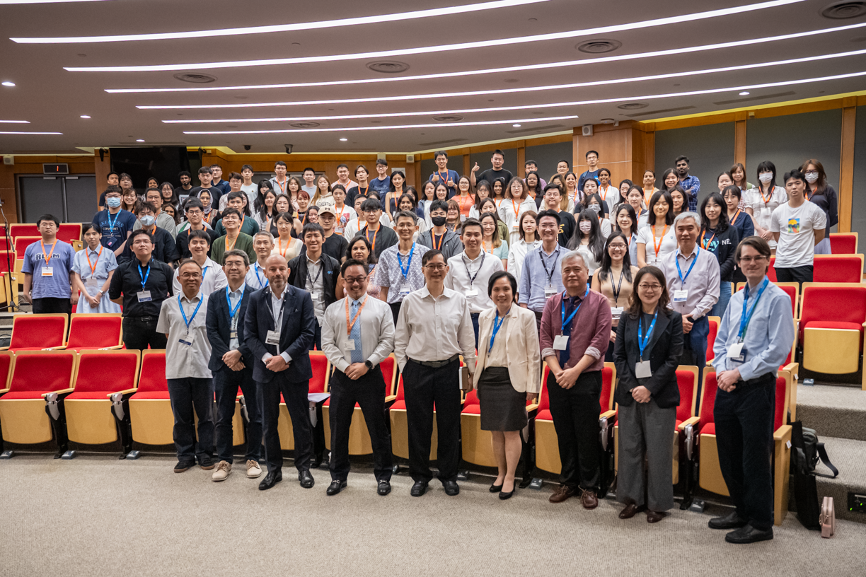 A large group of people wearing badges and conference lanyards pose together in an auditorium with red seats.